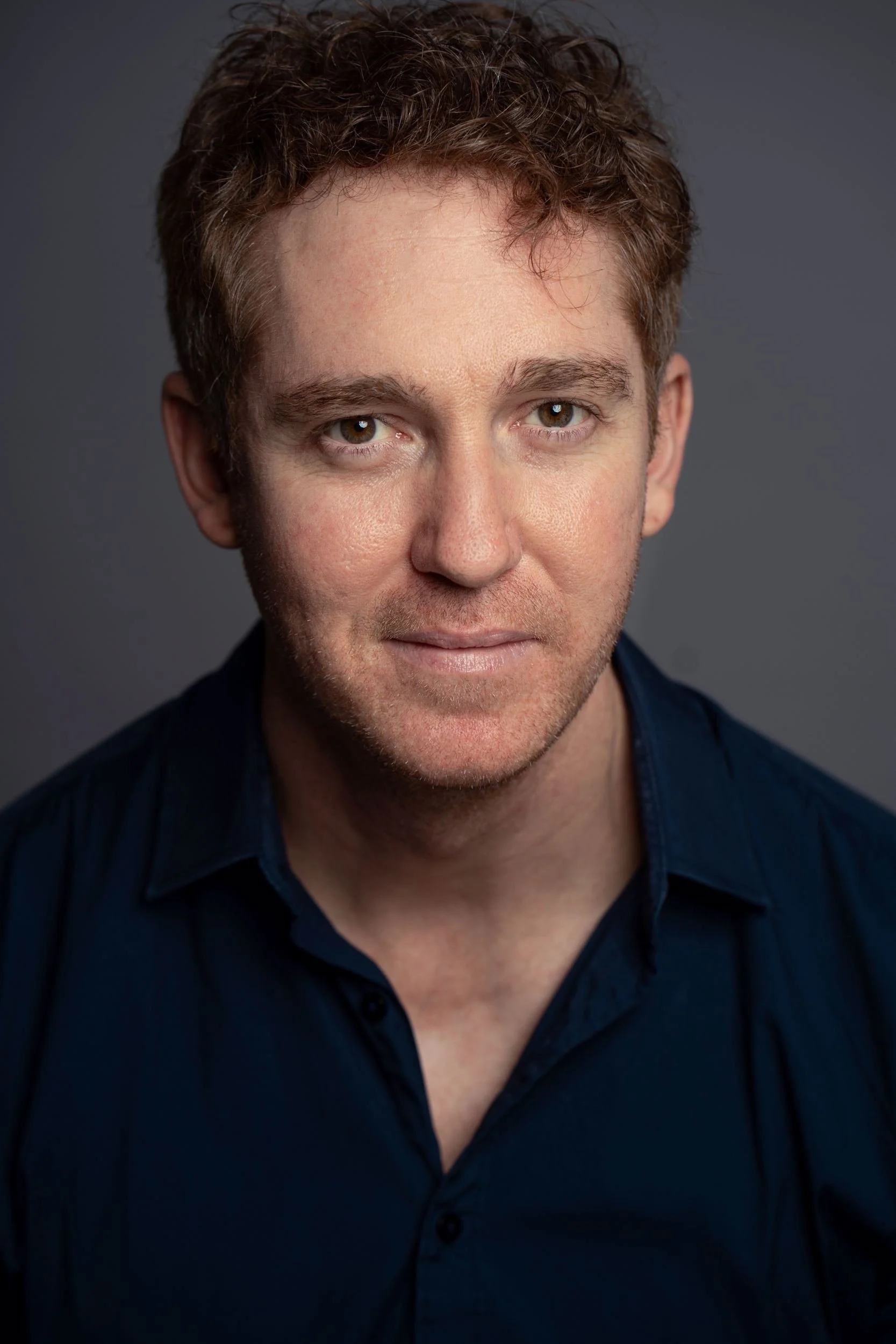 Close-up of a man with curly brown hair, hazel eyes, and light skin, wearing a dark blue shirt against a plain gray background.