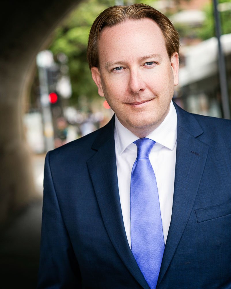 A man in a navy suit and light blue tie standing outdoors, smiling at the camera with a city street in the background.