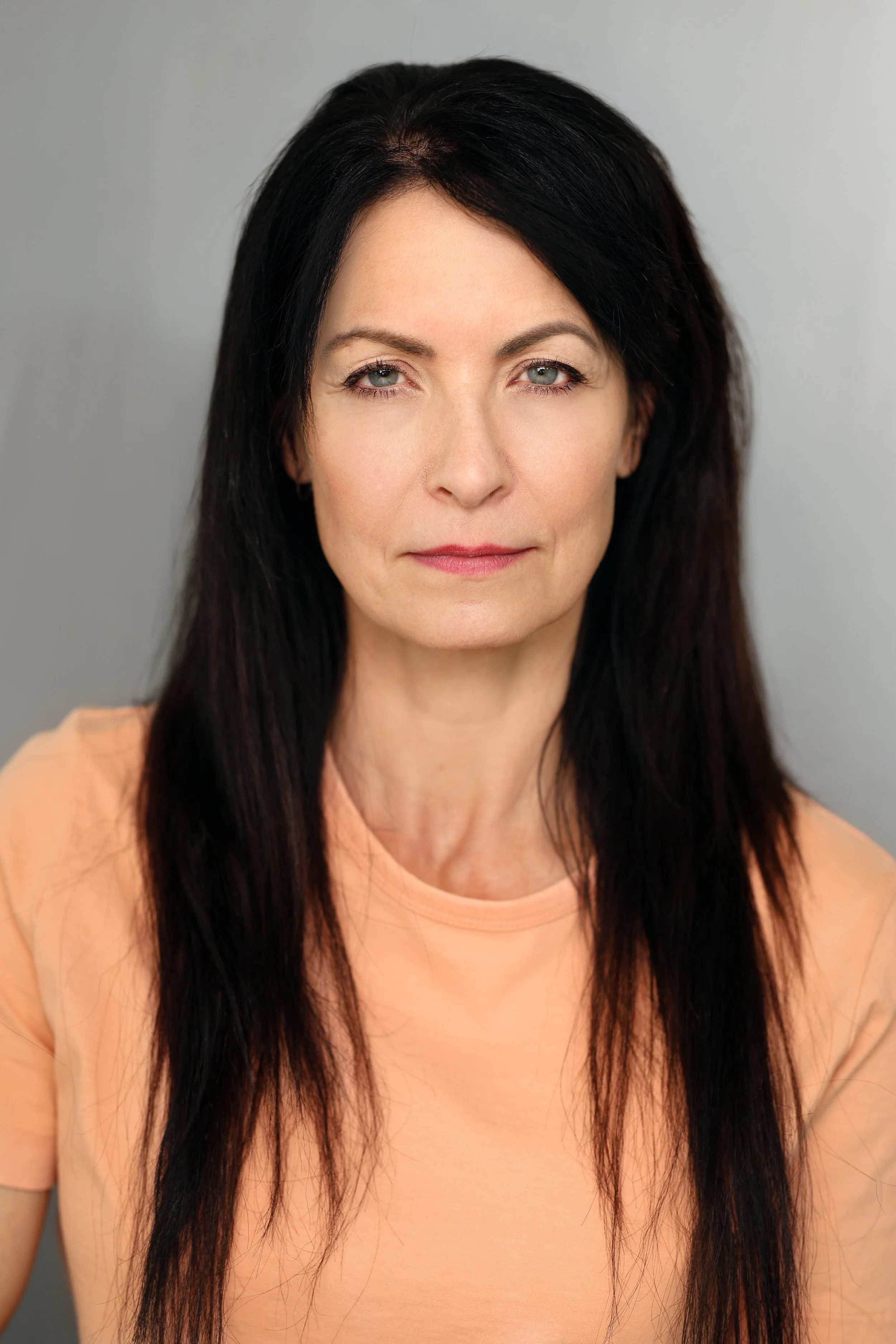 A woman with long black hair and light skin wearing a peach-colored top, looking directly at the camera with a neutral expression.