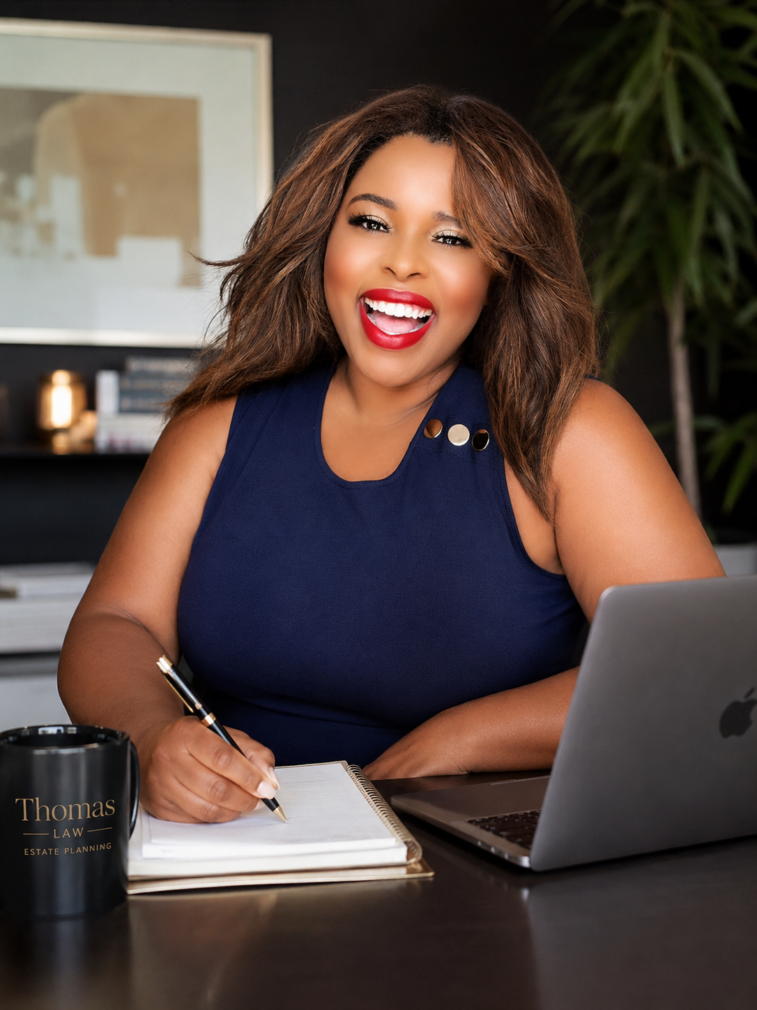 A smiling woman with long wavy hair, wearing a navy sleeveless top with metallic accents, sitting at a desk with a laptop, notepad, and a black mug that says 'Thomas Law Estate Planning' in gold lettering, in modern office setting.