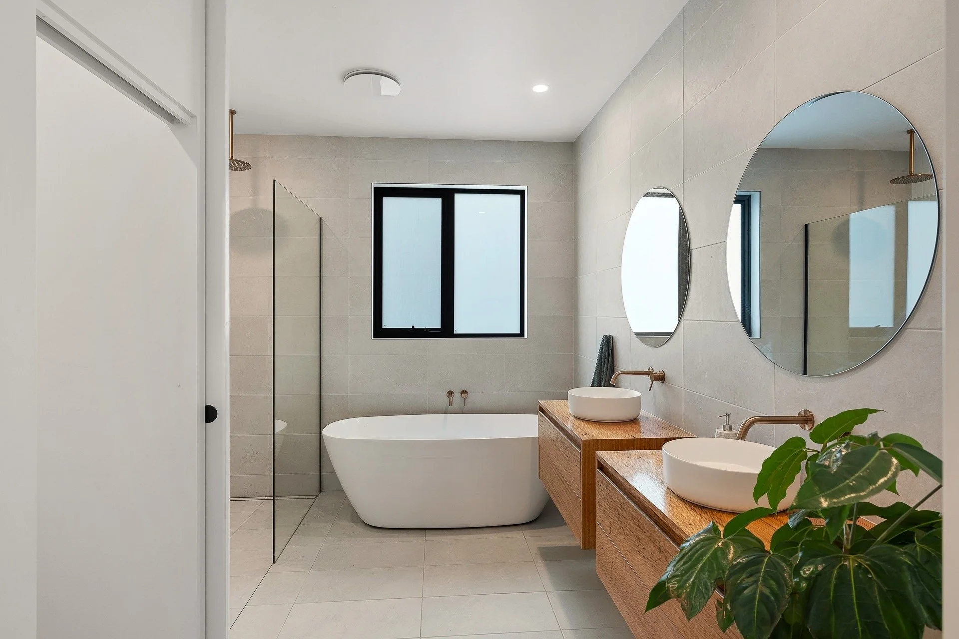 Modern bathroom with a white freestanding bathtub, two round vessel sinks on a wooden vanity, large round mirrors, a black-framed window, and a potted plant in the foreground.