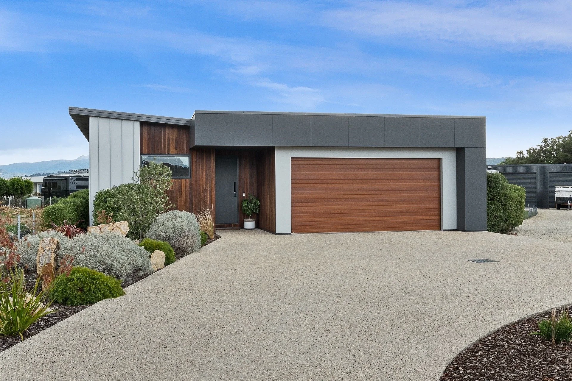 Modern house with a wooden garage door, dark front door, and a small front garden with various plants, shrubs, and rocks. Clear sky with a few clouds.