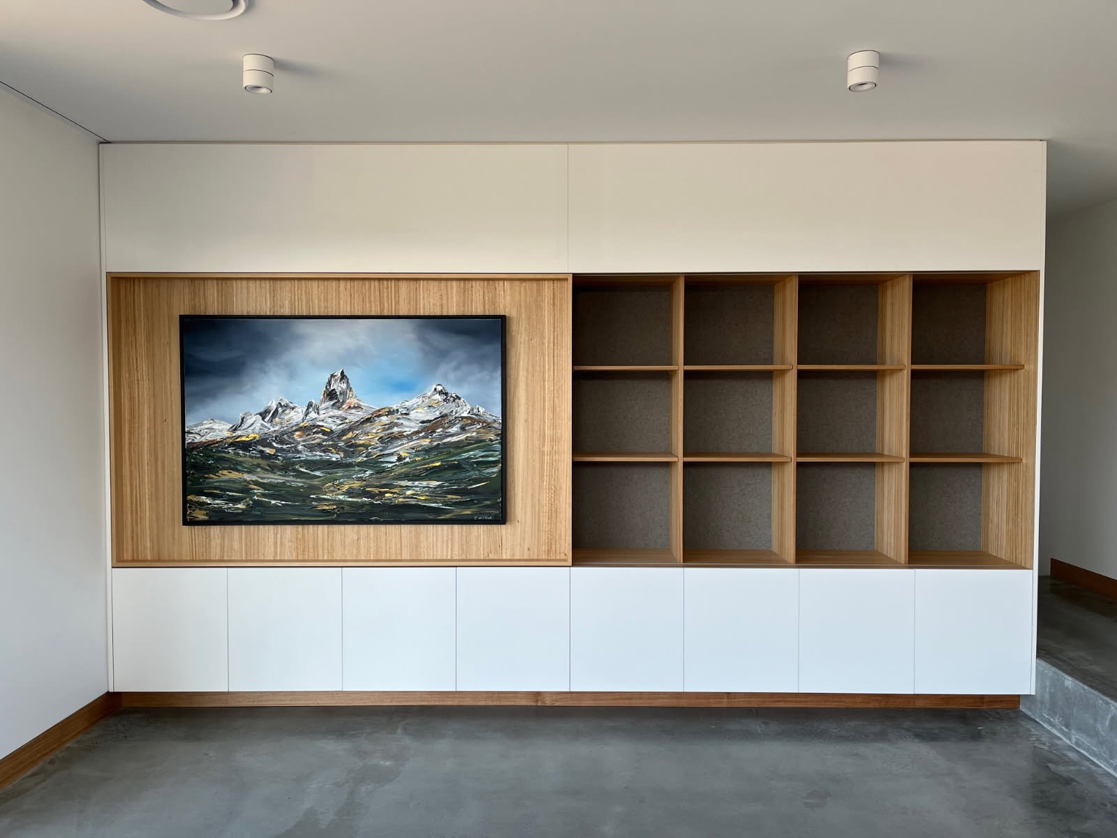 Interior view of a modern living room with a built-in wooden and white wall unit. The wall unit features a large framed painting of mountains with a cloudy sky and a section with empty shelves.