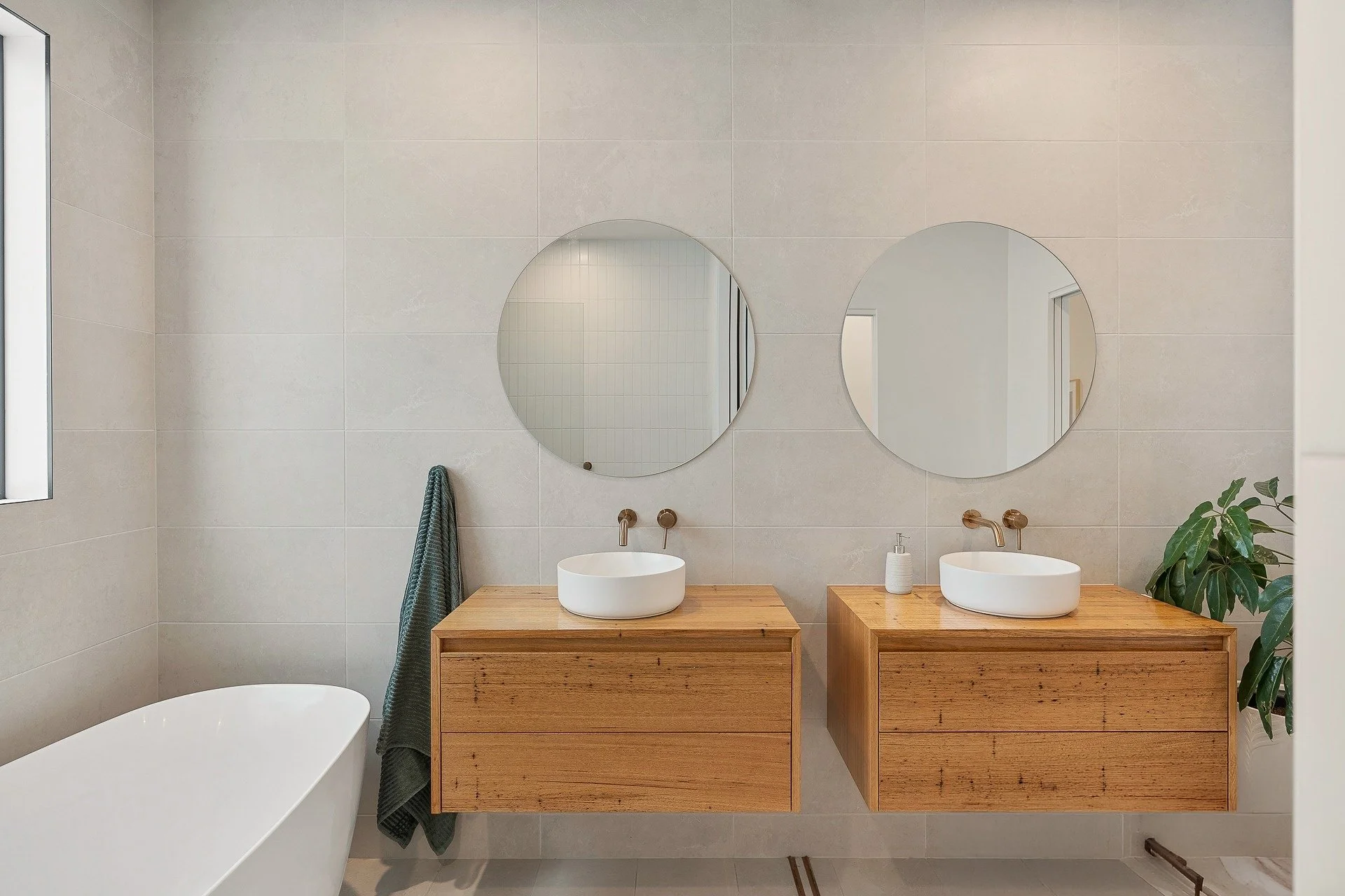 Bathroom vanity area with two oval mirrors, two vessel sinks on wooden cabinets, a faucet, a soap dispenser, a towel hanging on a hook, and a plant in the corner.