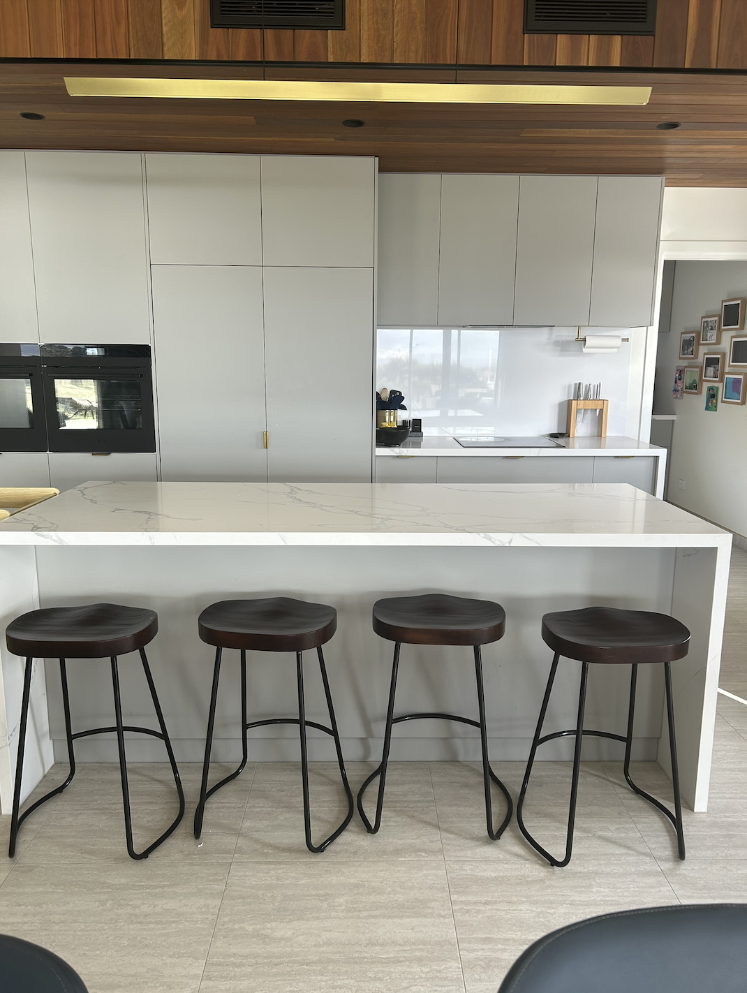 Modern kitchen with white marble island, four black bar stools, white cabinets, wooden accents, and a window with a view outside.