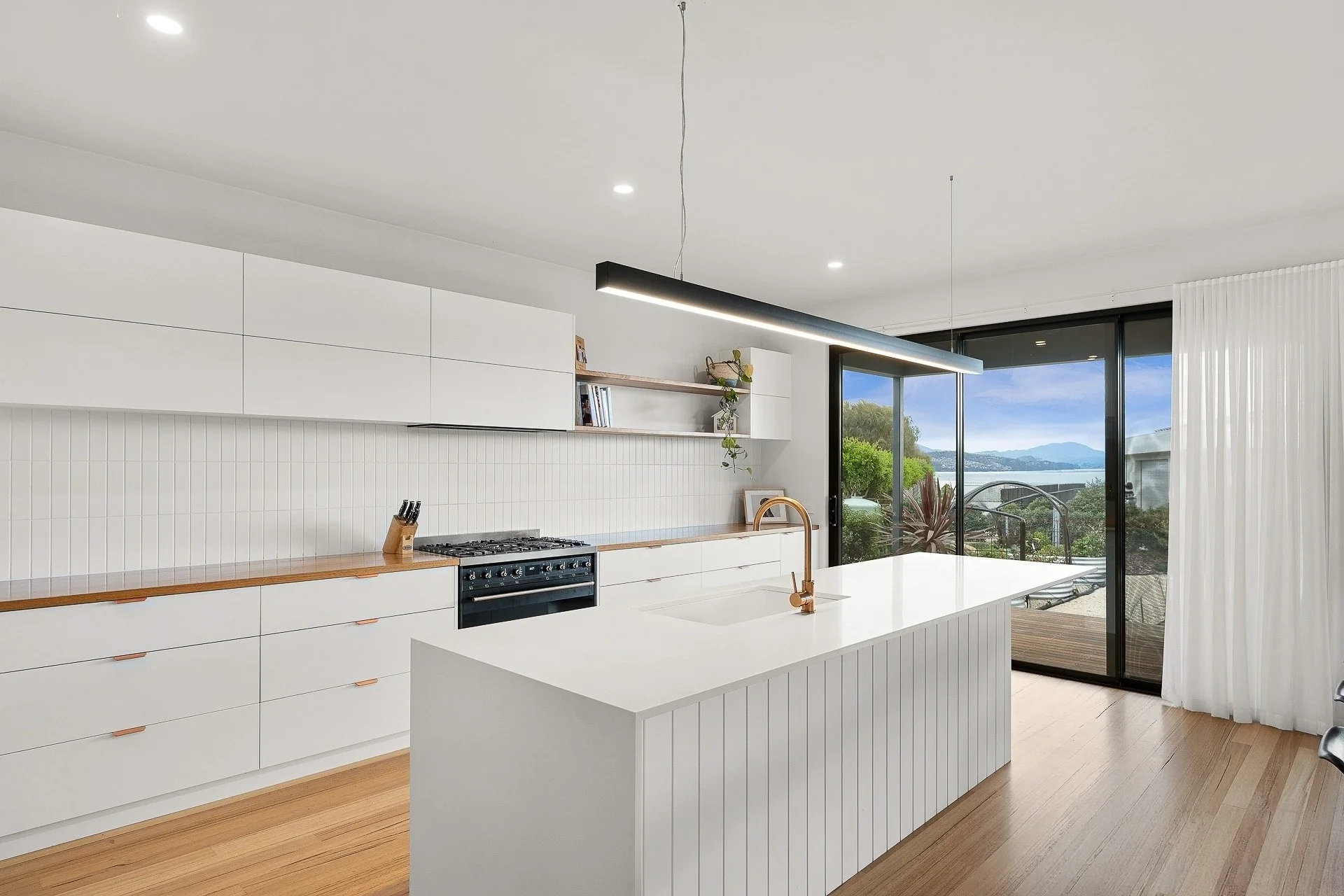 Modern white kitchen with a central island, built-in stove, wooden countertops, and large sliding glass doors overlooking a balcony and scenic water view.