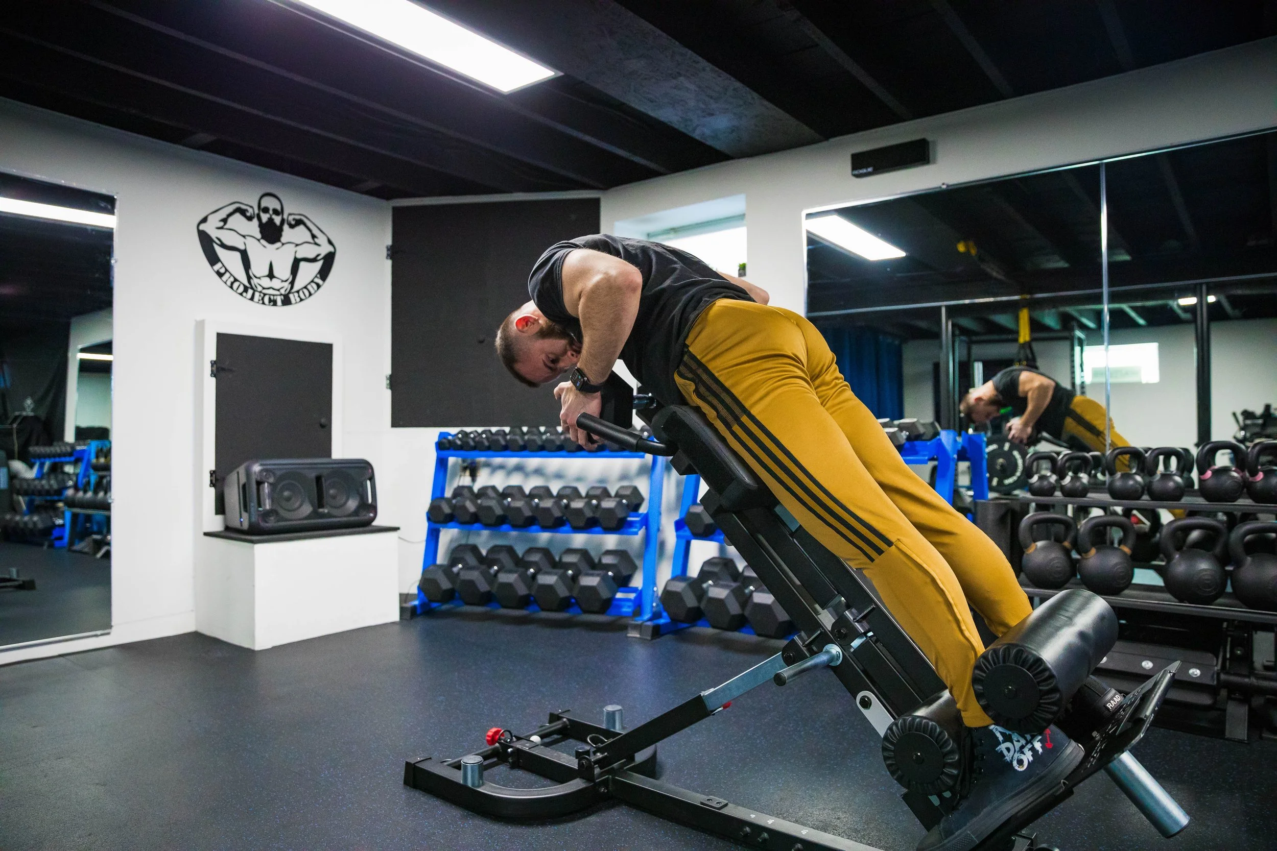 A man in black shirt and yellow pants is performing a hyperextension exercise on a hyperextension bench inside a gym. He is bent forward with his hands behind his head, working his lower back muscles. There are dumbbells and kettlebells visible on ra