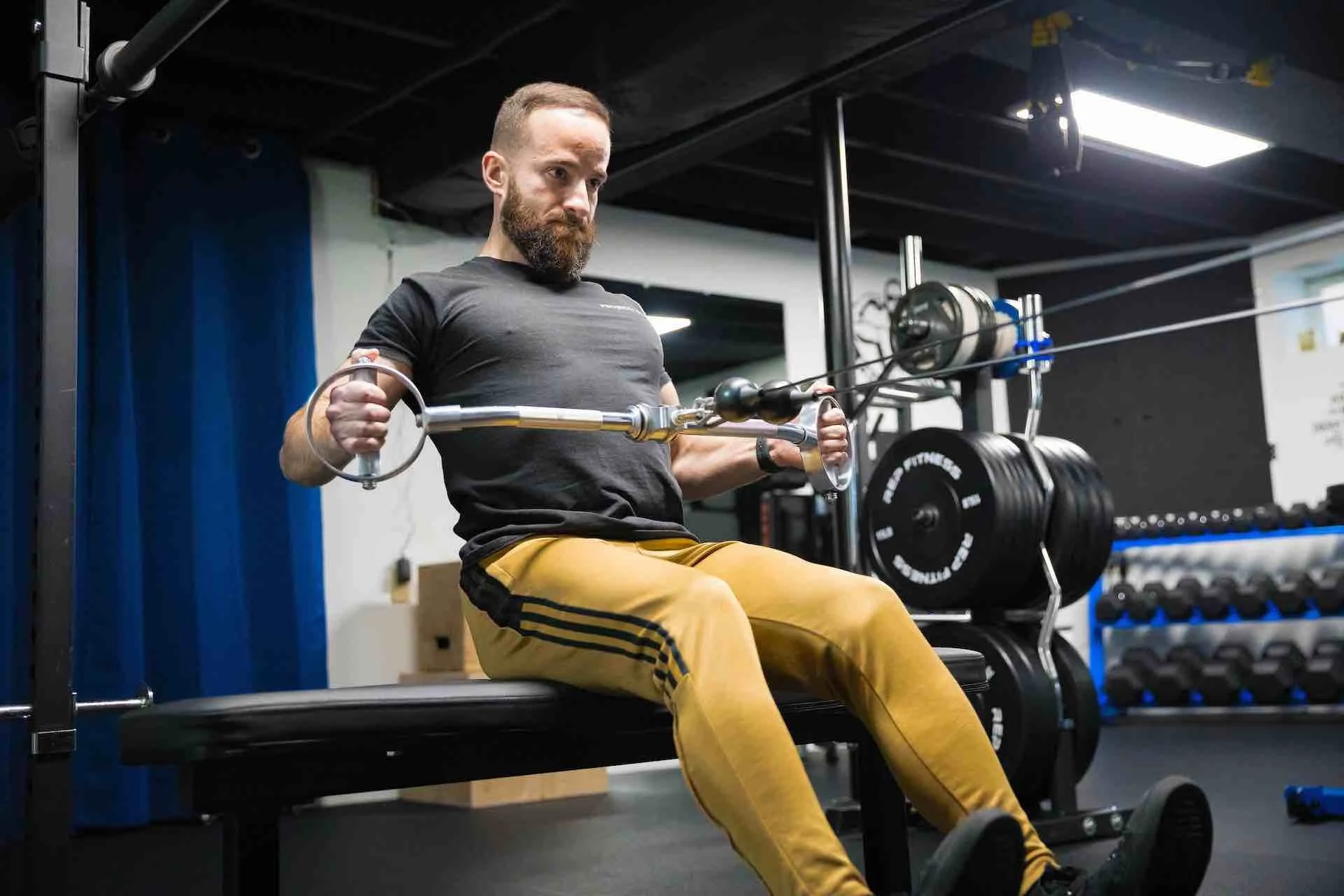 Man with beard in black T-shirt and yellow pants performing seated cable row exercise at gym.