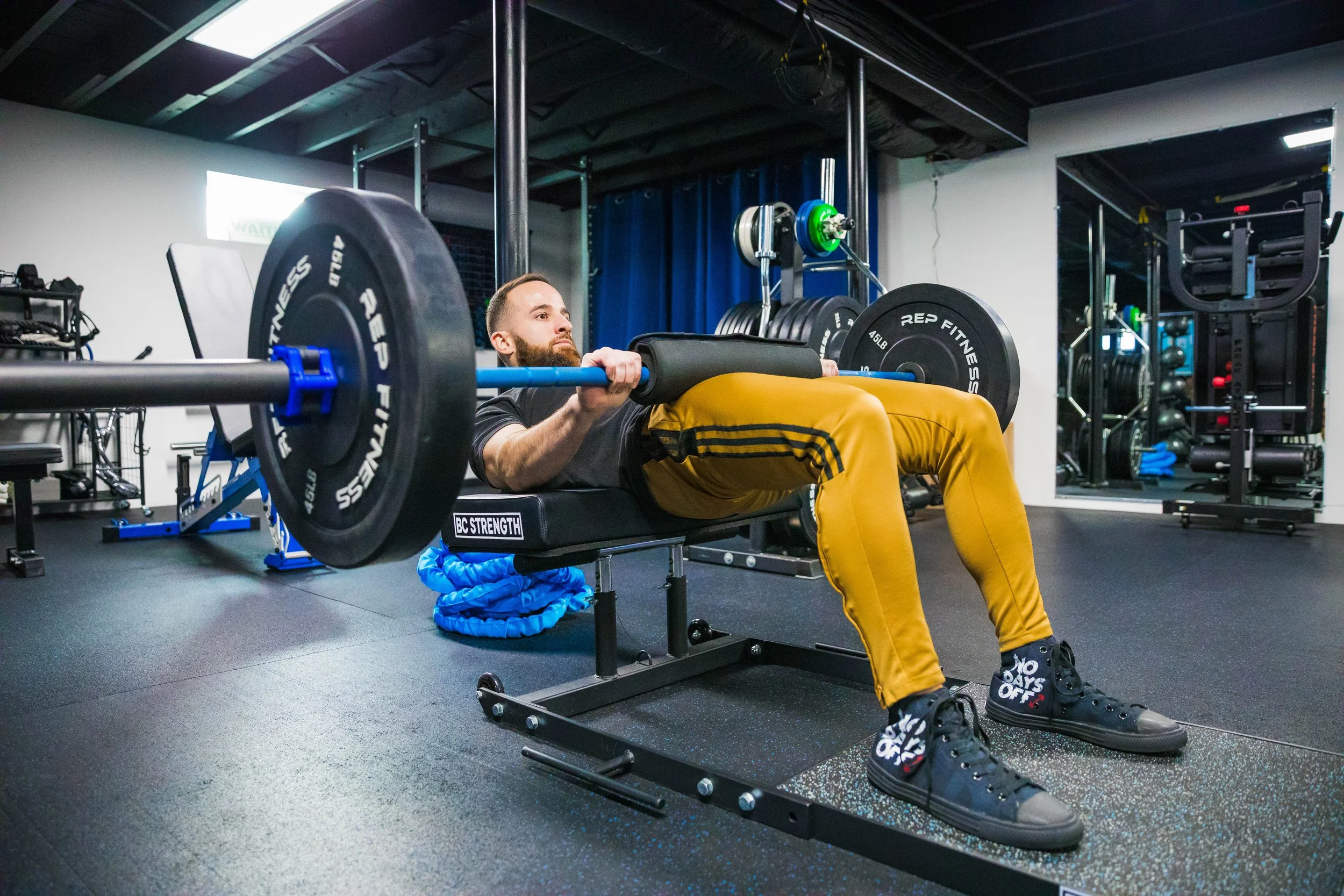 A man is lying on a workout bench in a gym, lifting a barbell with weight plates, surrounded by exercise equipment.