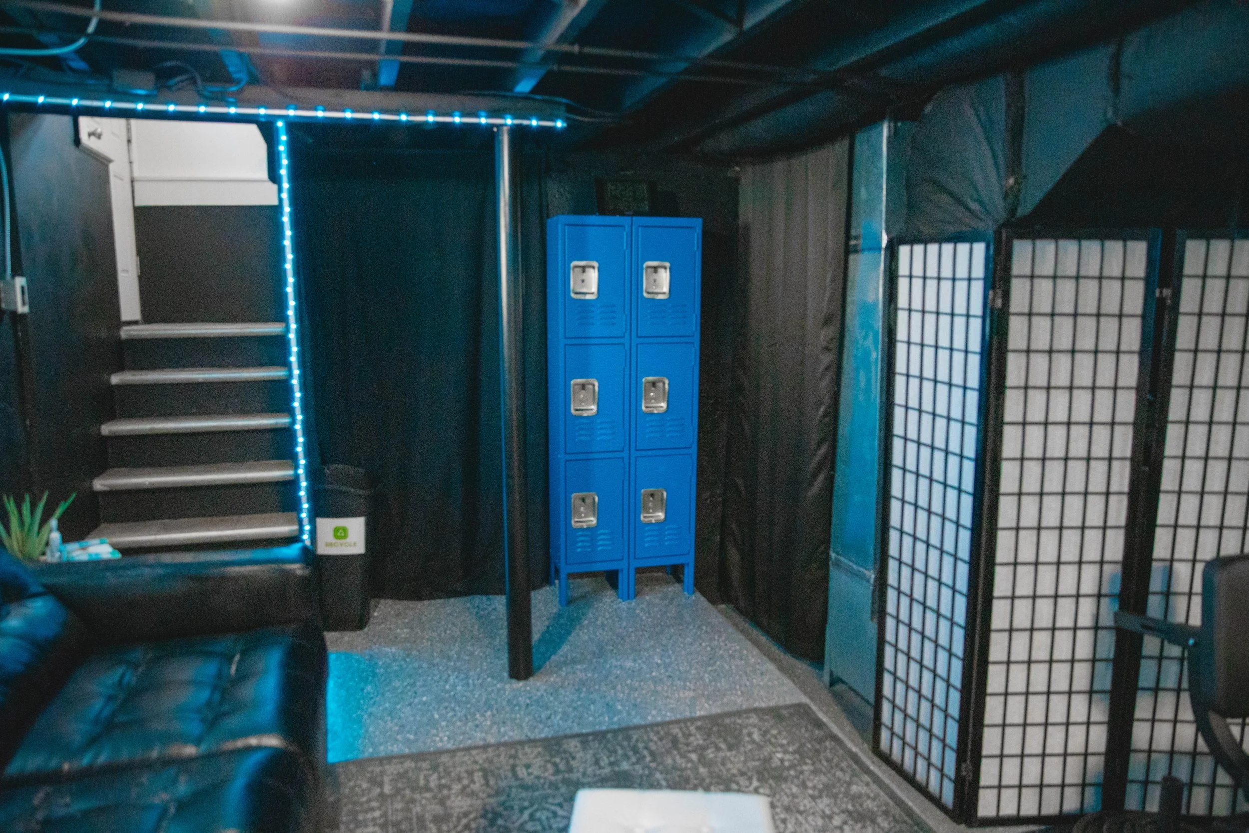 Indoor space with black walls and ceiling, featuring metal stairs with white and gray steps, bright blue lockers, a black leather sofa partially visible, and a folding room divider with black wooden frame and white paper panels, illuminated by blue s