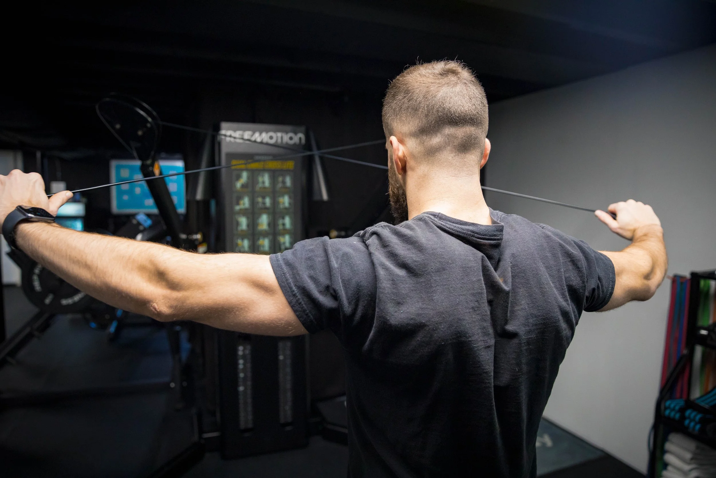 A man with a beard and short hair, wearing a black t-shirt, is doing lateral raises with a resistance band in a gym.