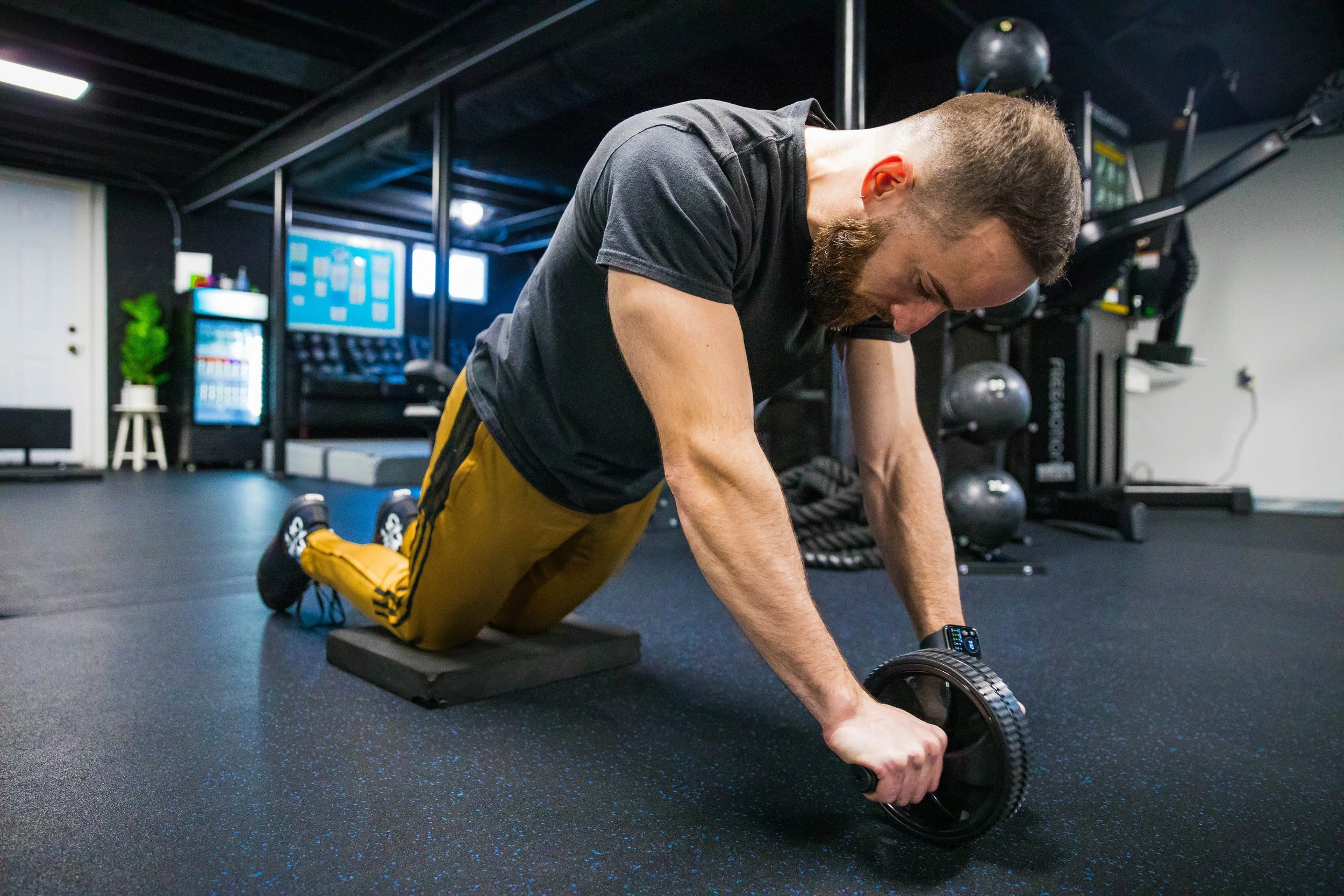 Man in a black T-shirt and yellow pants performing a workout with an ab roller on a black mat in a gym.