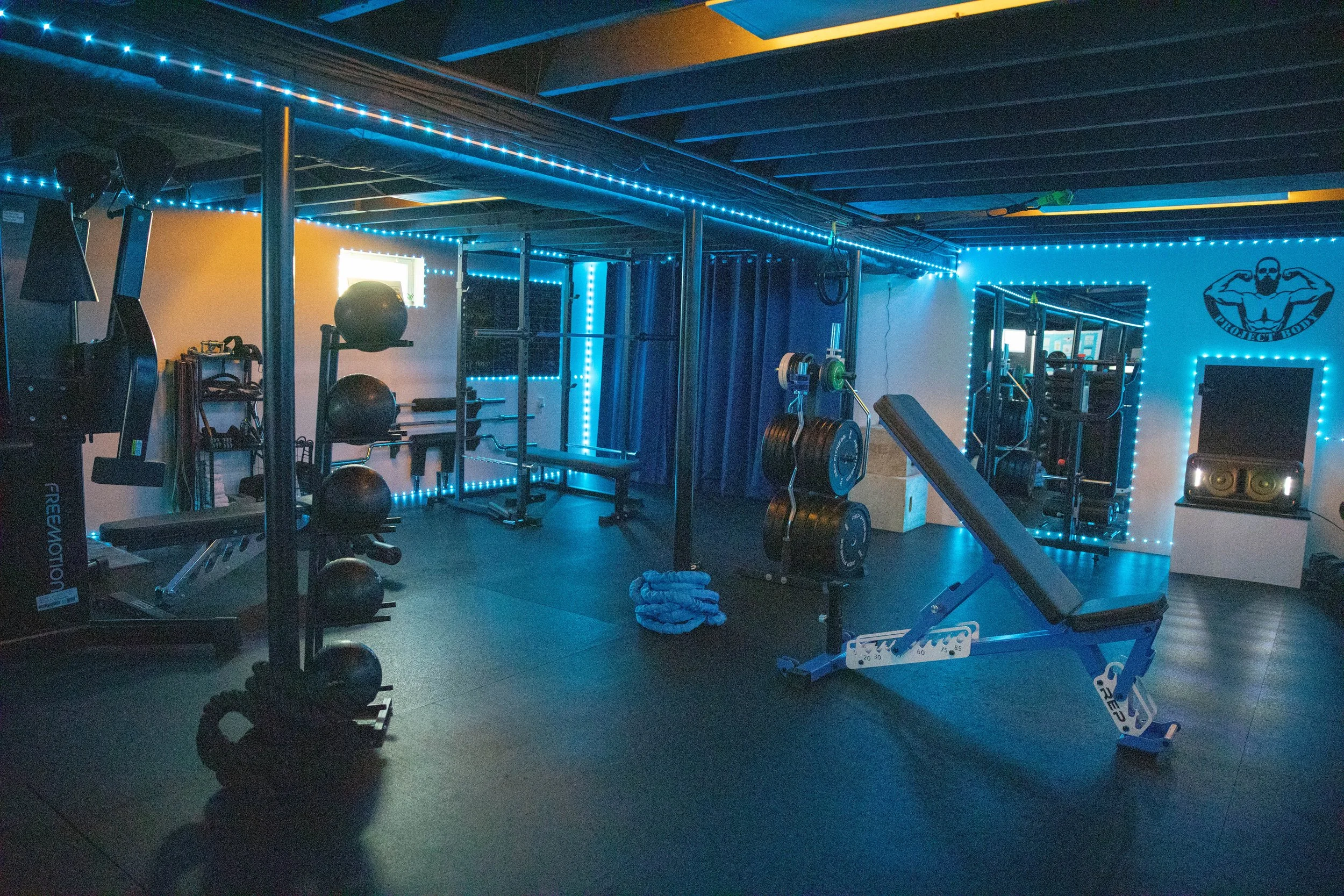 Home gym with various workout equipment, blue LED strip lights, a large mirror, and a logo on the wall of a muscular man flexing.