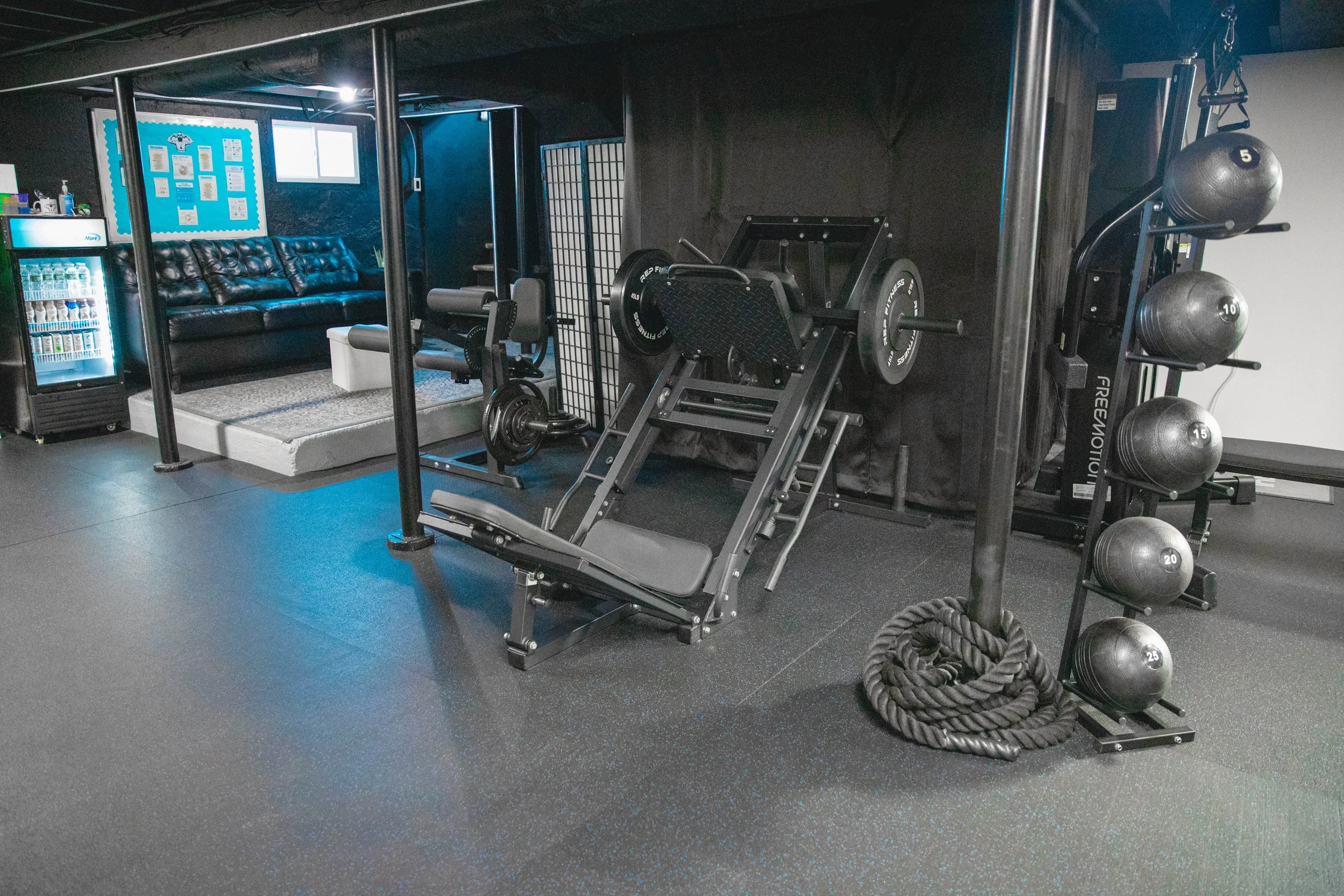 Gym area with weightlifting equipment, including a leg press machine, a rack of medicine balls, and a battle rope, with a seating area and beverage fridge in the background.