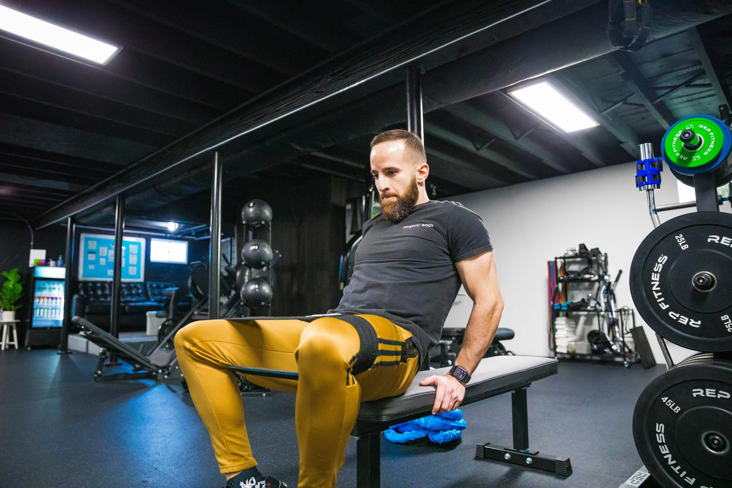 A man with a beard in a black t-shirt and yellow pants performing a strength exercise on a bench in a gym, with fitness equipment and weights around him.