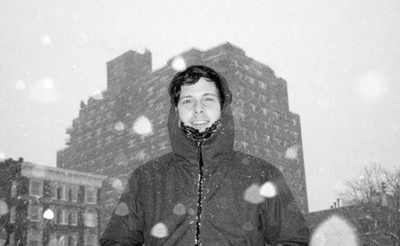 A man in a winter jacket standing outside in snowy weather with falling snowflakes and tall buildings in the background.