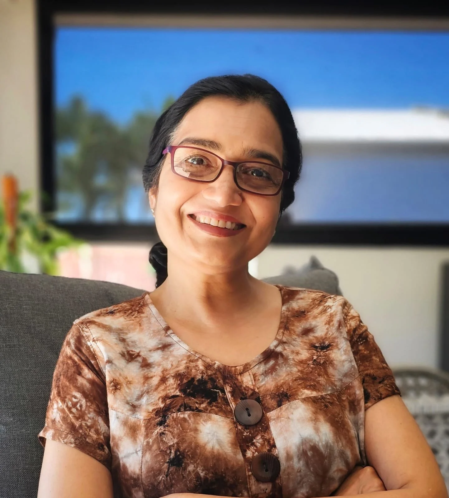 A woman with short black hair, glasses, and a brown patterned shirt smiling and sitting in a chair indoors with a TV and plants in the background.