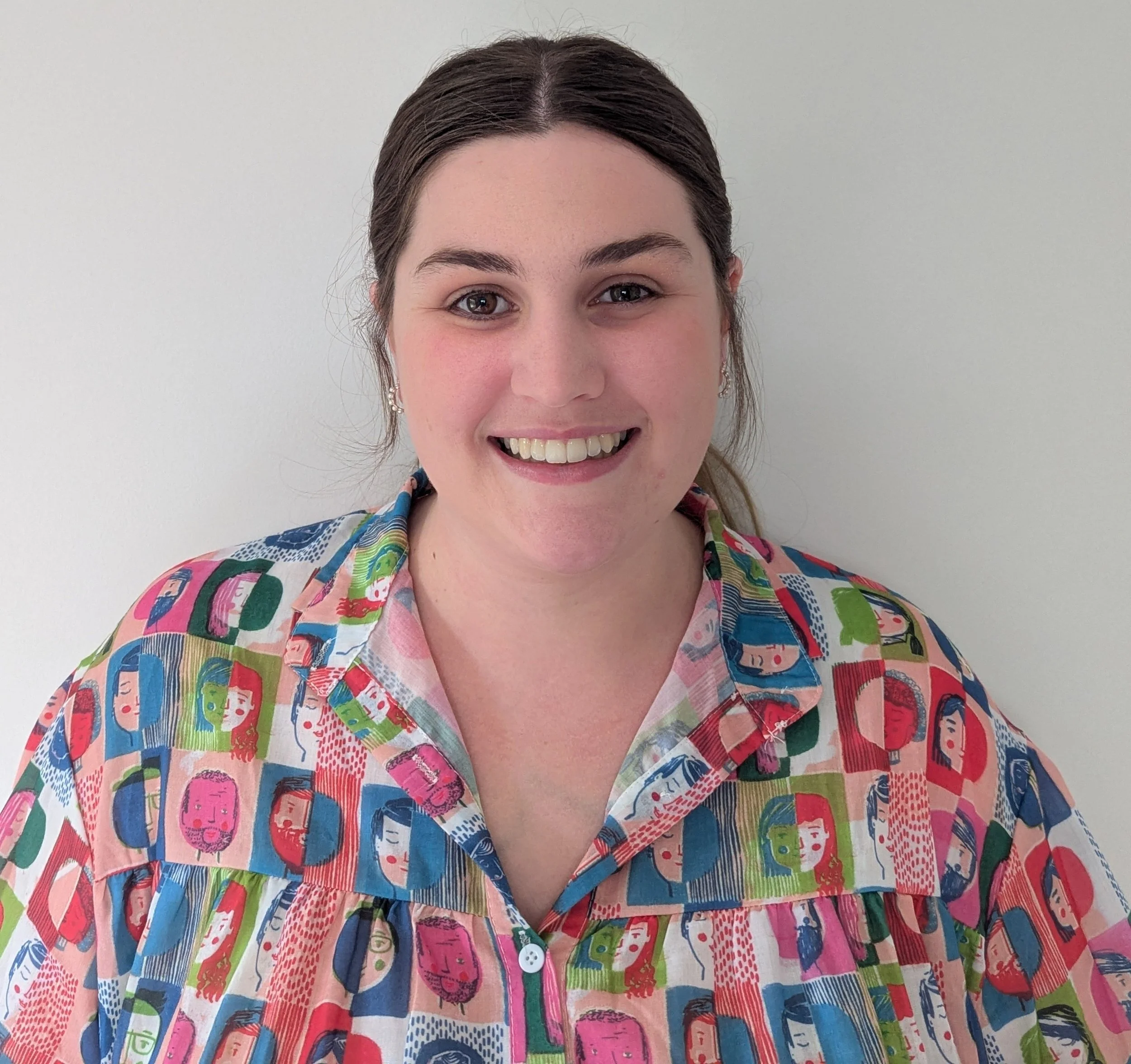 A young woman with dark brown hair smiling at the camera against a plain white wall. She is wearing a colorful, patterned button-up shirt with various illustrated faces.