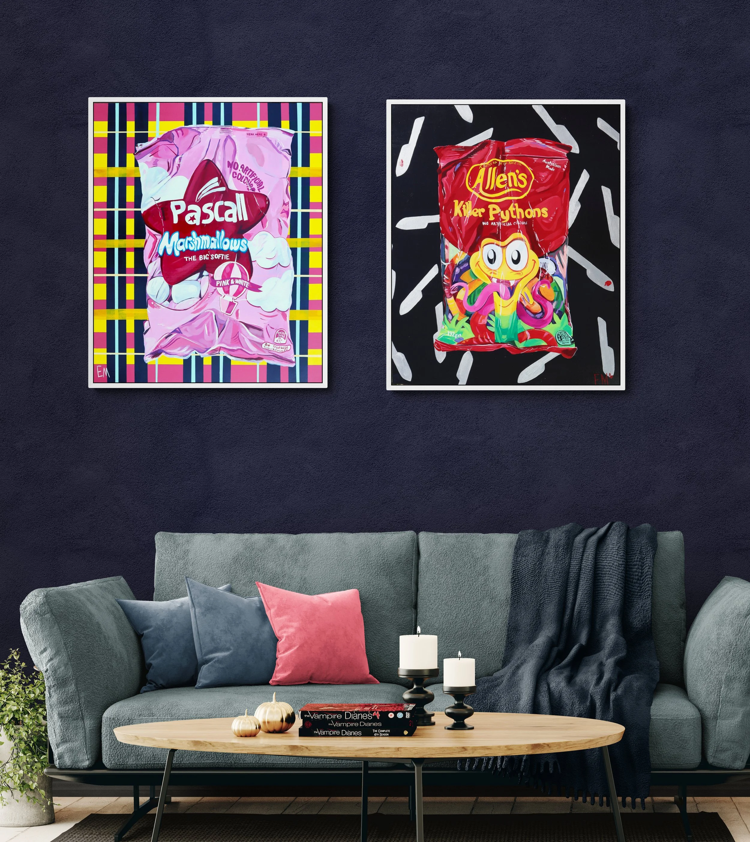 Two colorful art posters of candy bags hanging on a dark wall above a gray couch with pillows and a wooden coffee table with candles and small pumpkins.