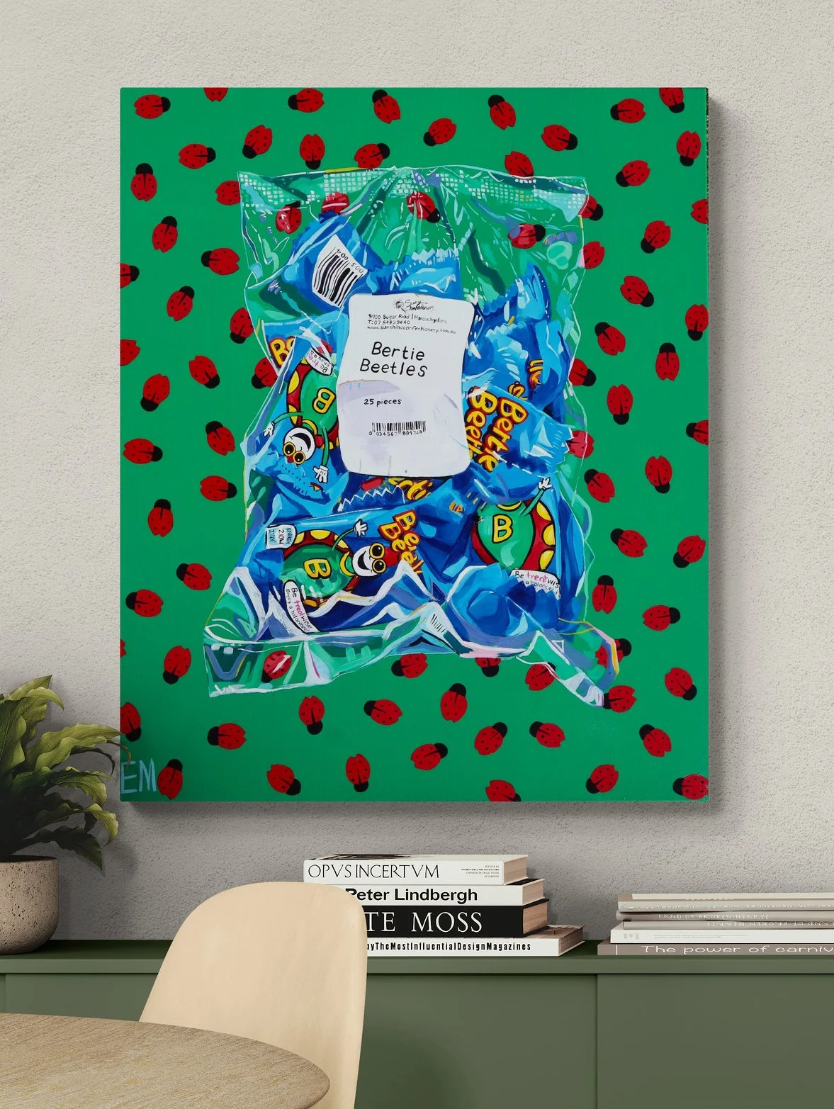 A colorful wall art piece featuring a green background with scattered red ladybugs. Overlaid on the wall art, there is a plastic bag containing multiple Bertie Beetle chocolates with labels, placed on the wall. In front, there is a beige chair and a green shelf with stacked books.