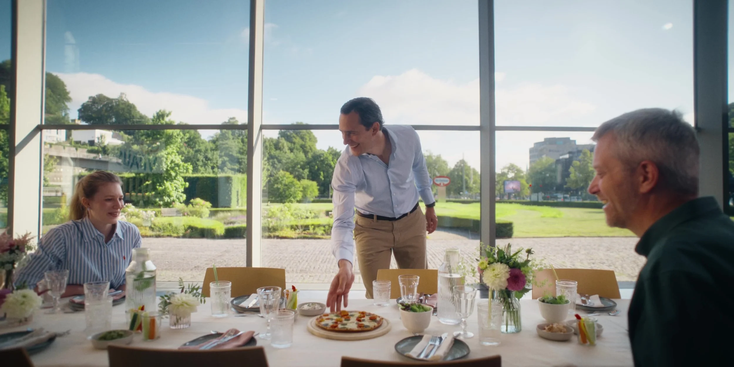 People sitting at a table in a restaurant with large windows showing greenery outside. A man in a white shirt is serving or reaching for a pizza on the table. A woman with a striped shirt and an older man are seated, smiling and engaging in the meal.