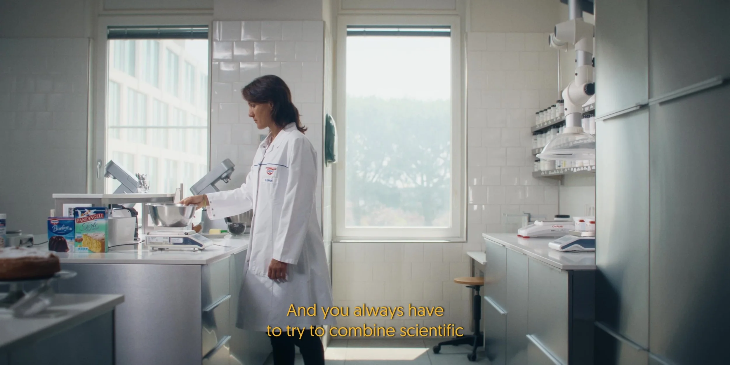 A scientist in a white lab coat working in a laboratory, standing at a counter with scientific equipment and food ingredients, with large windows in the background.