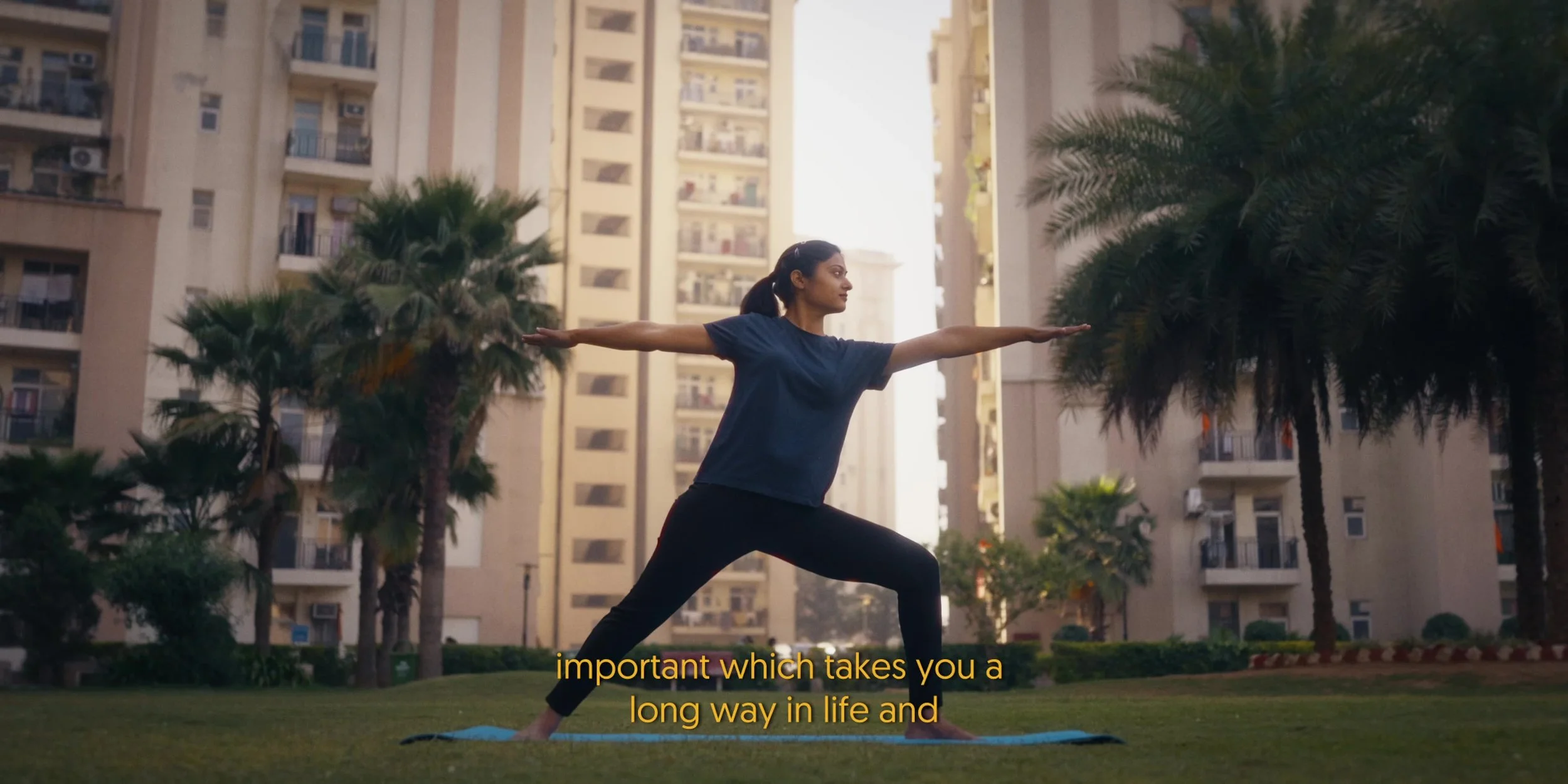A woman practicing yoga outdoors on a grassy area surrounded by palm trees and tall apartment buildings, with subtitles reading 'important which takes you a long way in life and'.