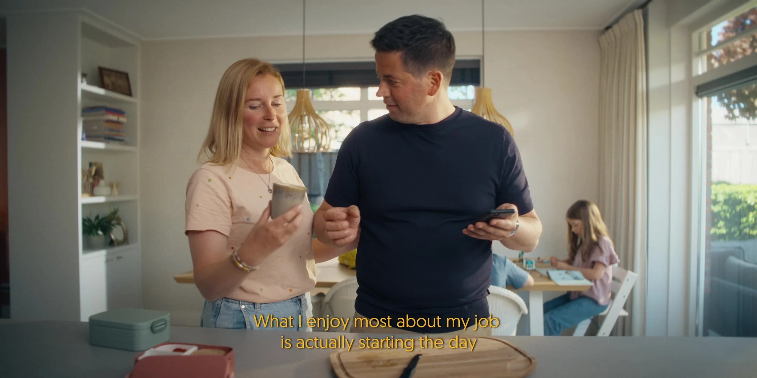 A man and woman standing in a kitchen, looking at a phone together, with the woman smiling. Two children are sitting at the table in the background, engaged in an activity. Subtitles read, 'What I enjoy most about my job is actually starting the day.