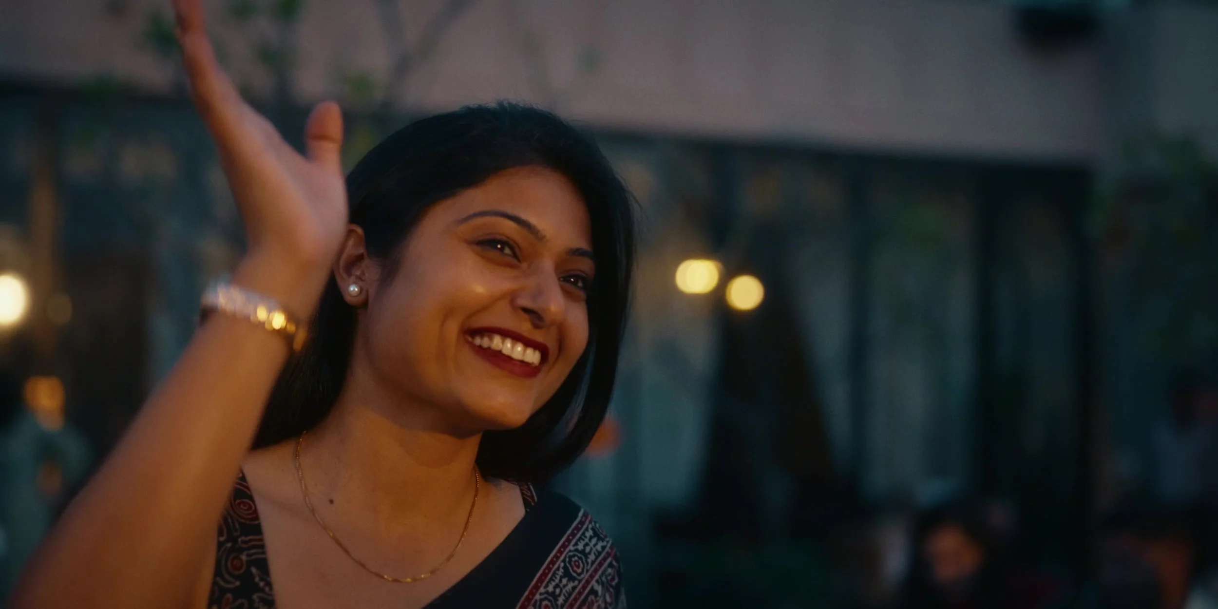 A woman with dark hair smiling and waving, wearing a patterned top and pearl earrings in an outdoor setting at dusk.