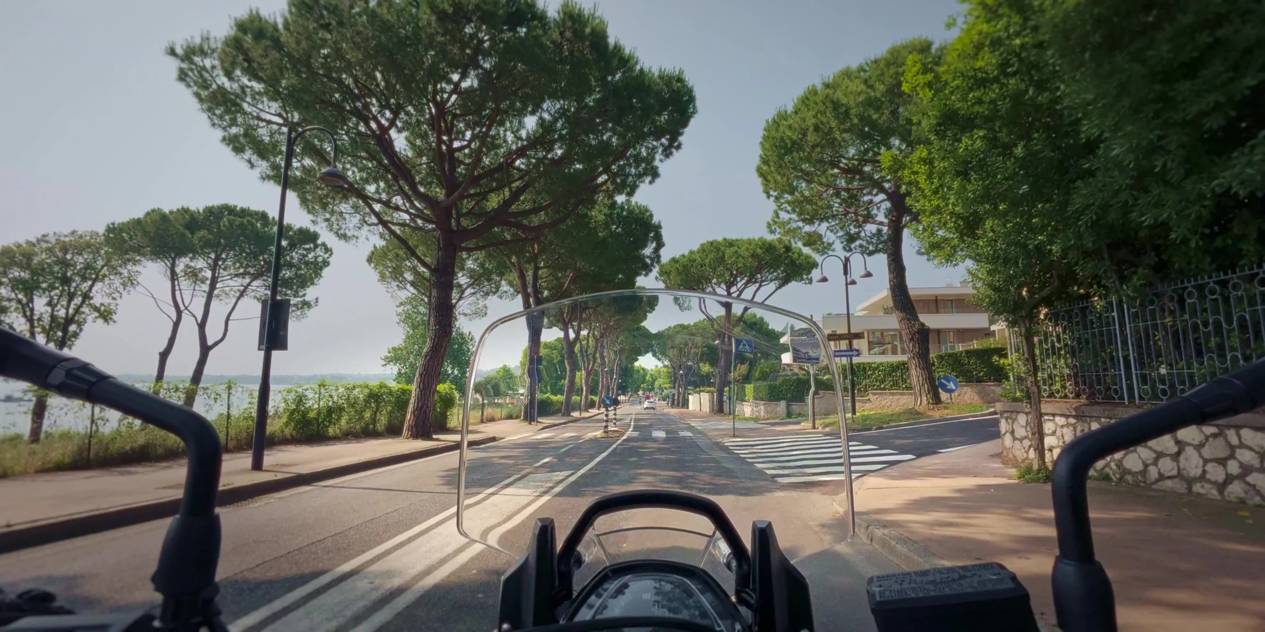 View from a motorcycle or bike on a tree-lined street, with a parking lane on the right, sidewalk, and residential buildings, under a clear sky.