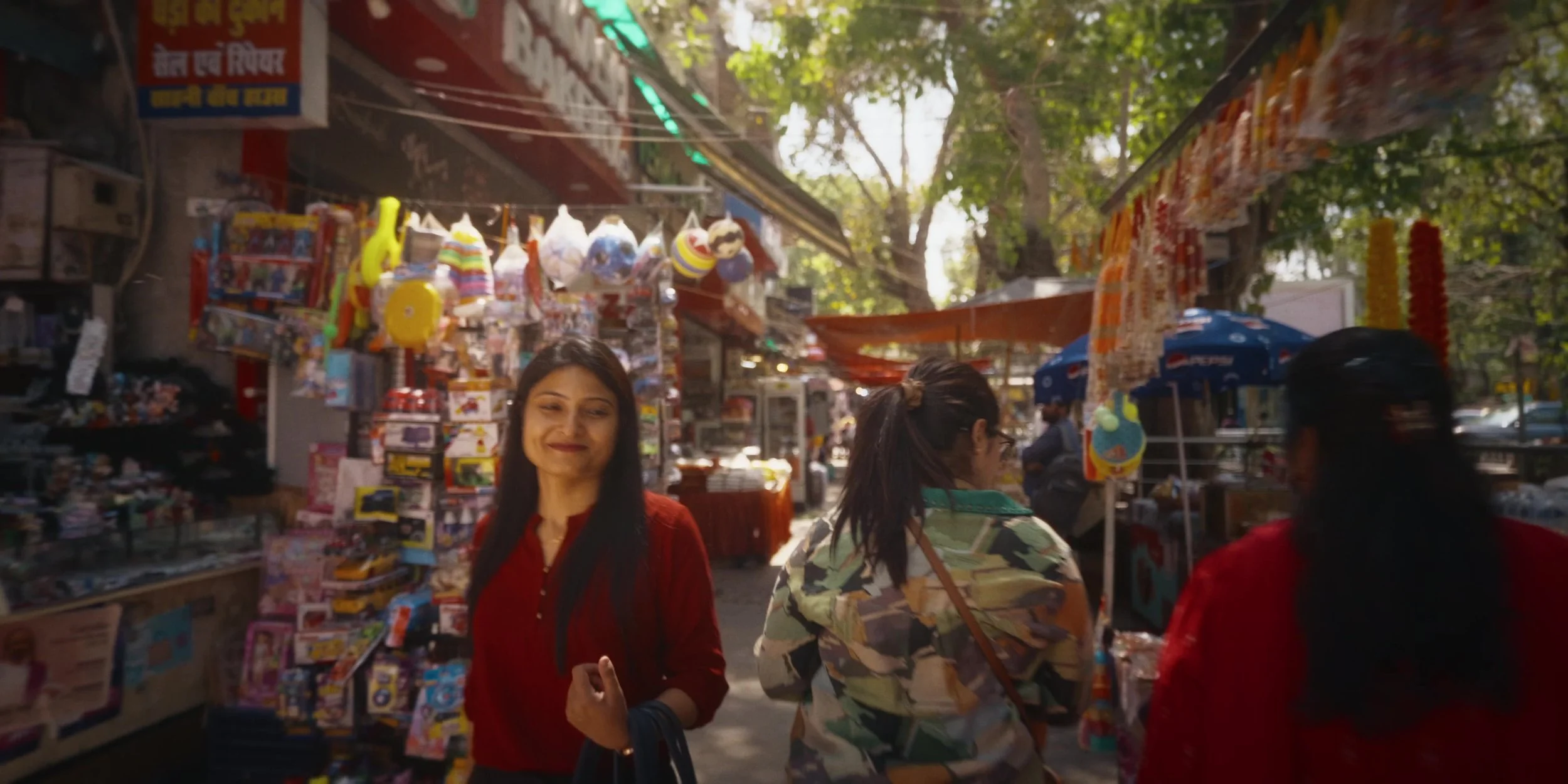 Women shopping at outdoor market stall with toys, balloons, and other items on display, under trees and sunlight.