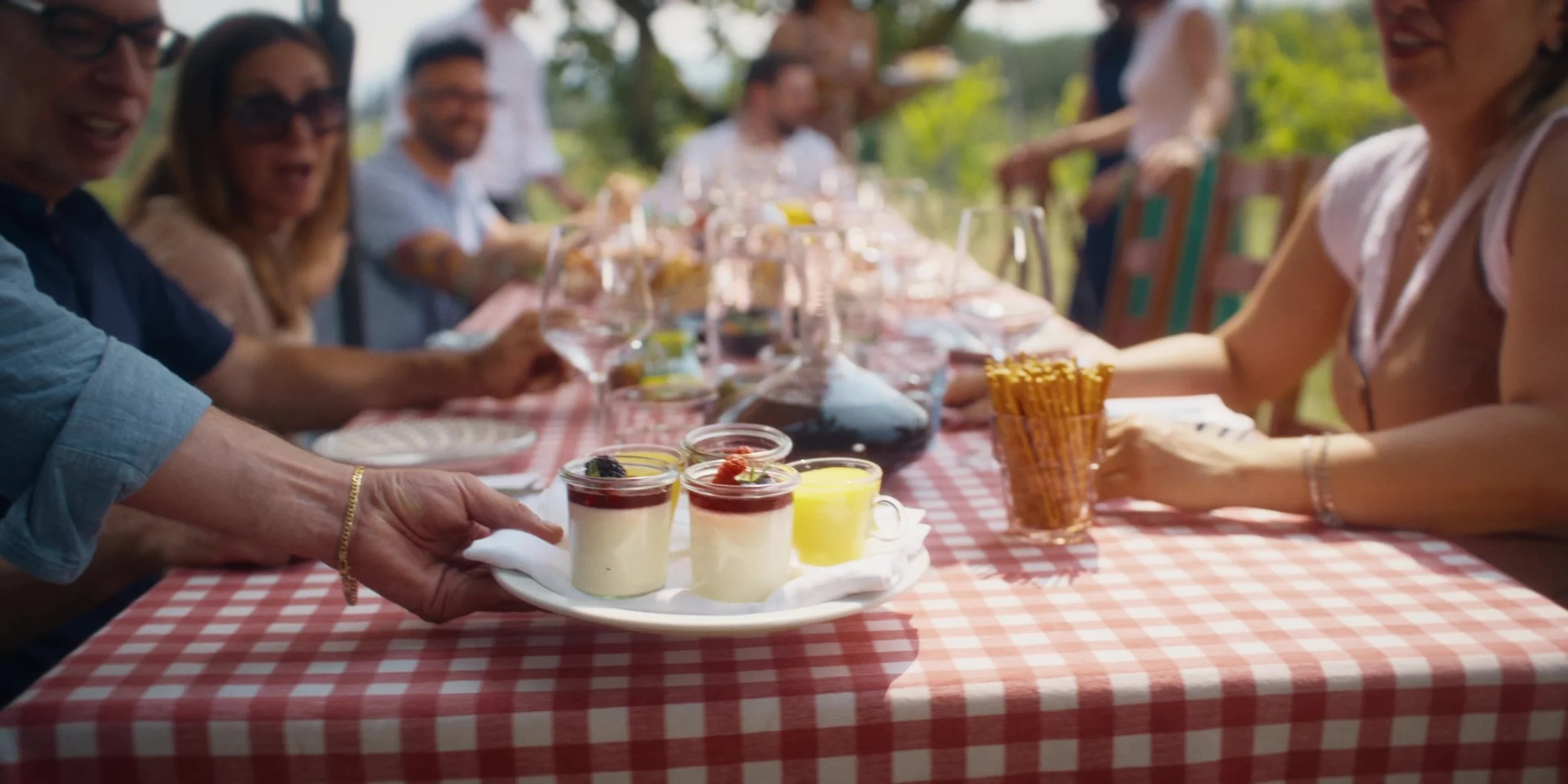 People gathered at a long outdoor table with a red checkered tablecloth, enjoying a meal with desserts, drinks, and appetizers during daytime.