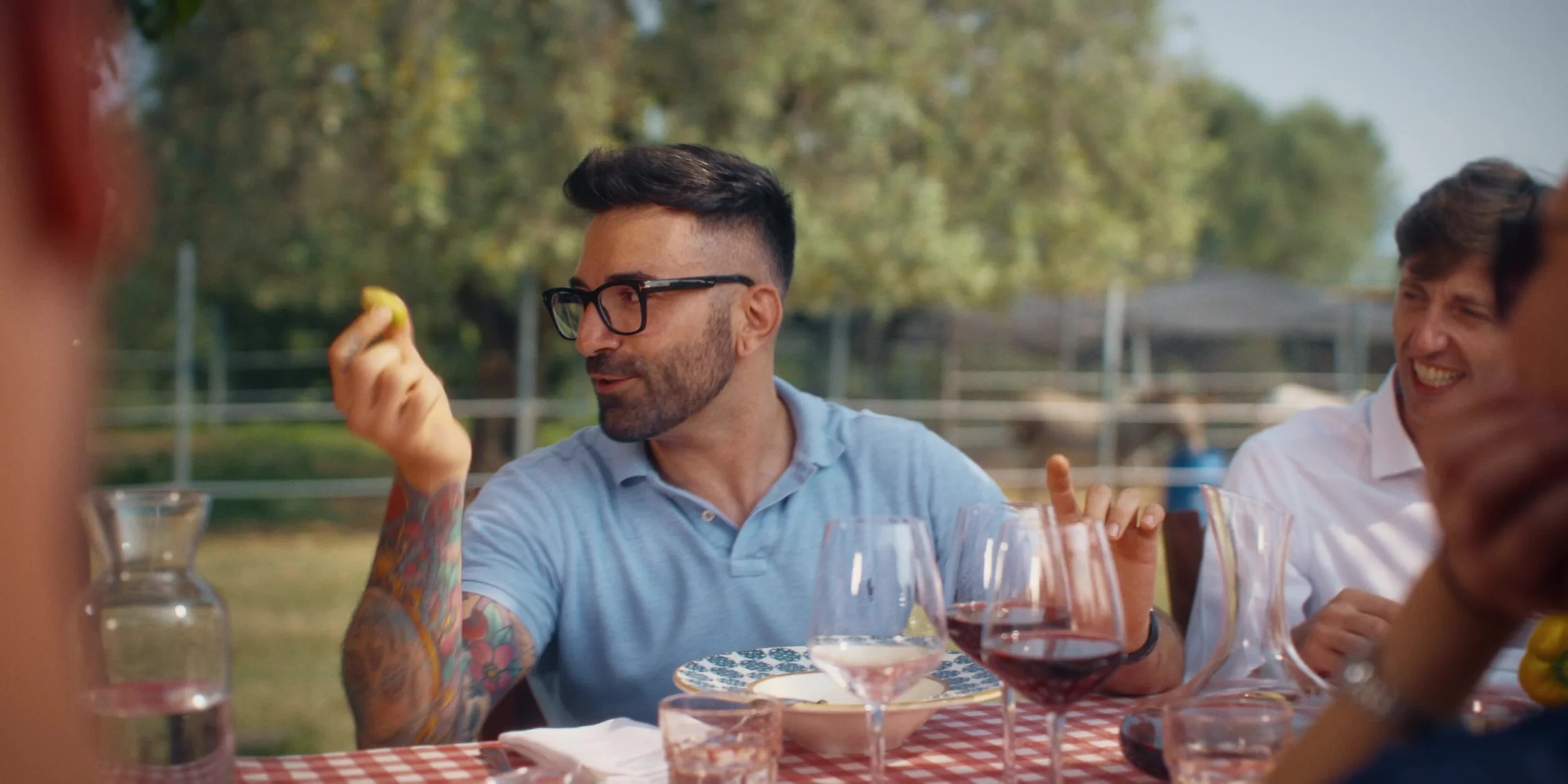 People enjoying a meal outdoors, with one man holding a lemon, surrounded by glasses of wine and water.