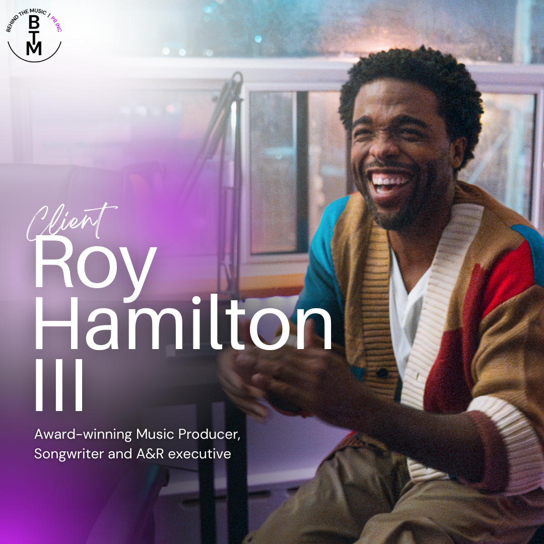 A man with dark curly hair and a beard, smiling and laughing, sitting in a room with large windows. He is wearing a multicolored jacket and a white shirt inside.