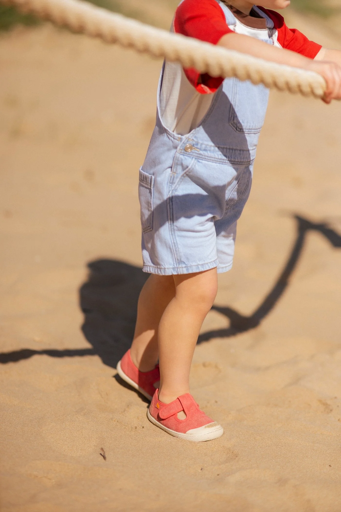 Niño vestido con camiseta roja y pantalones cortos de mezclilla, sosteniendo una cuerda en un entorno arenoso.