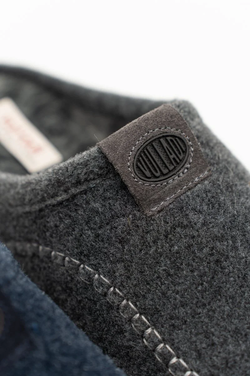 Detalle de la etiqueta de un zapato de tela gris con una costura visible y un logotipo ovalado en la parte superior.