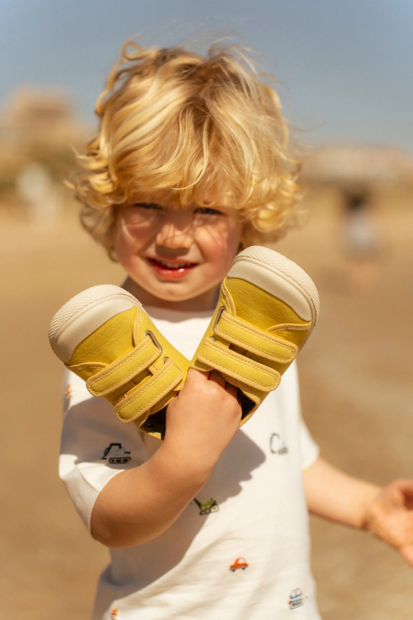 Niño rubio en la playa con guantes de boxeo amarillos, sonriendo y haciendo un gesto con el puño cerrado.