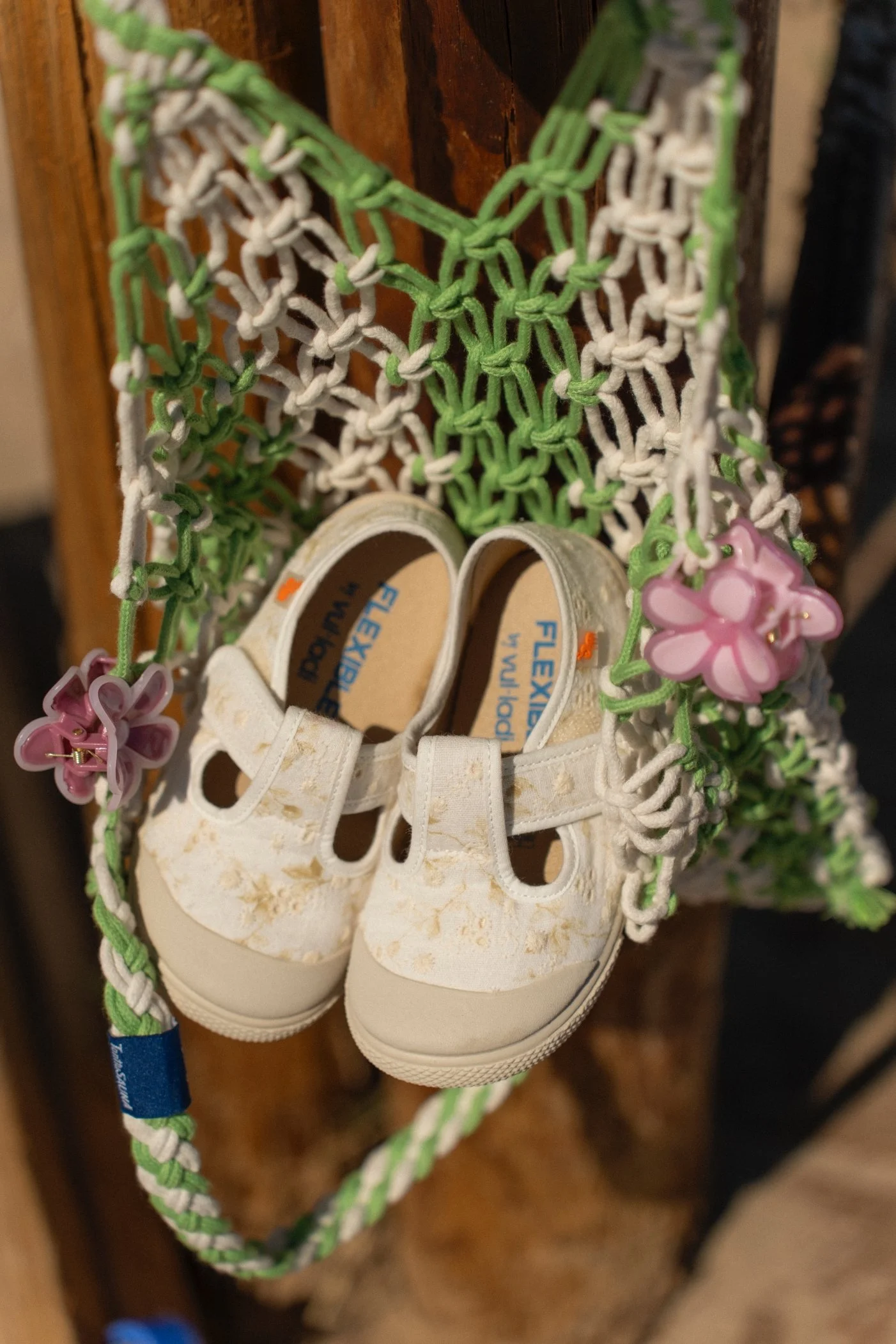 Un par de zapatos blancos pequeños con detalles dorados, colocados en una red de macramé verde y blanco, decorada con flores de plástico rosadas en un fondo de madera.
