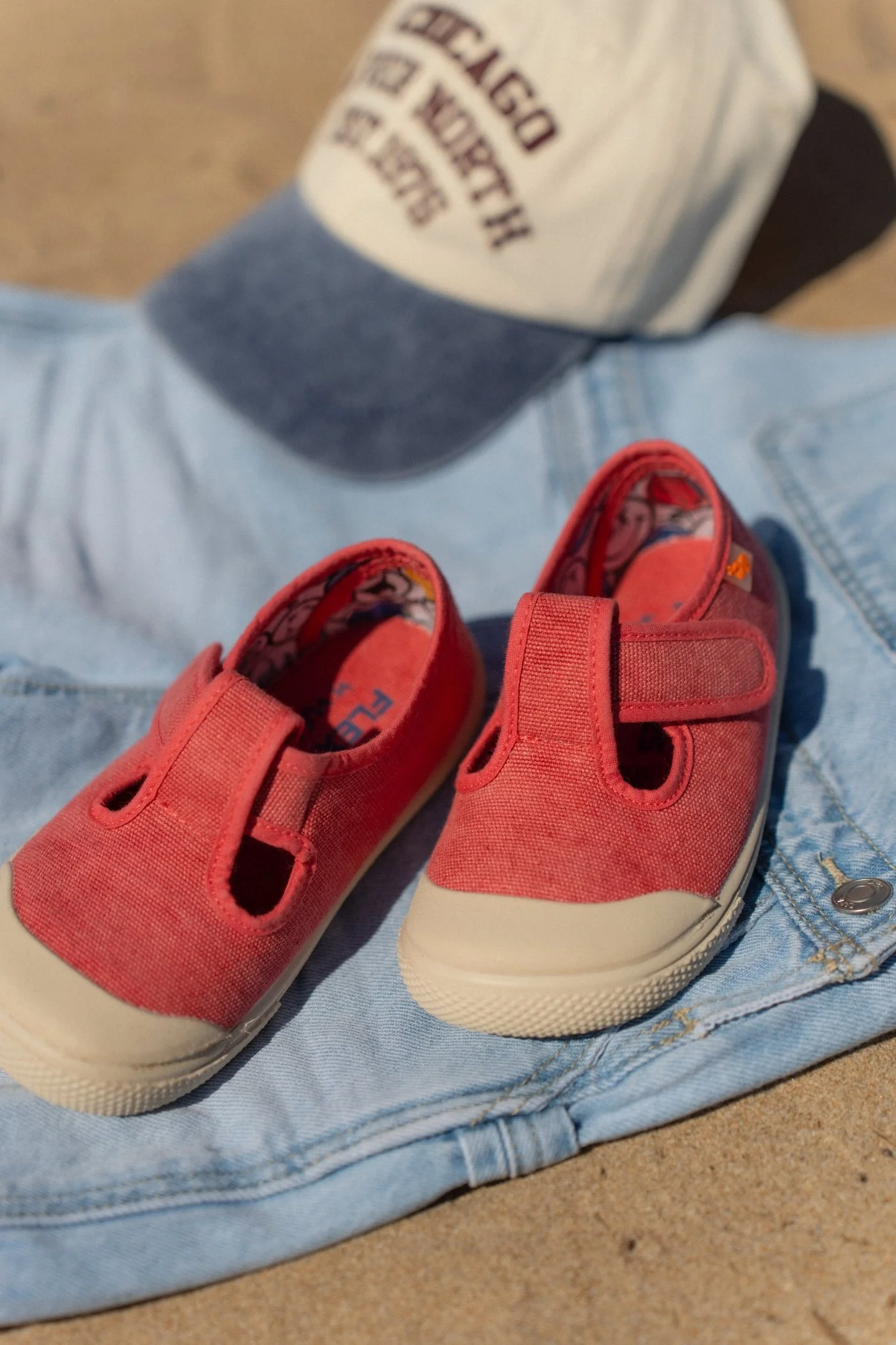 Un par de zapatos de mezclilla rojos con suela blanca, colocados sobre unos pantalones de mezclilla y junto a una gorra de béisbol. La gorra es de colores gris y blanco con letras en rojo. La escena está en una superficie de arena.