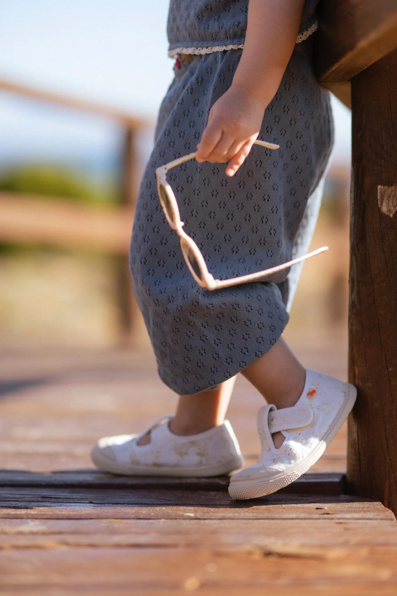 Niña con pantalones de mezclilla, sosteniendo gafas de sol, de pie en un muelle de madera, con un fondo difuso y cielo despejado.