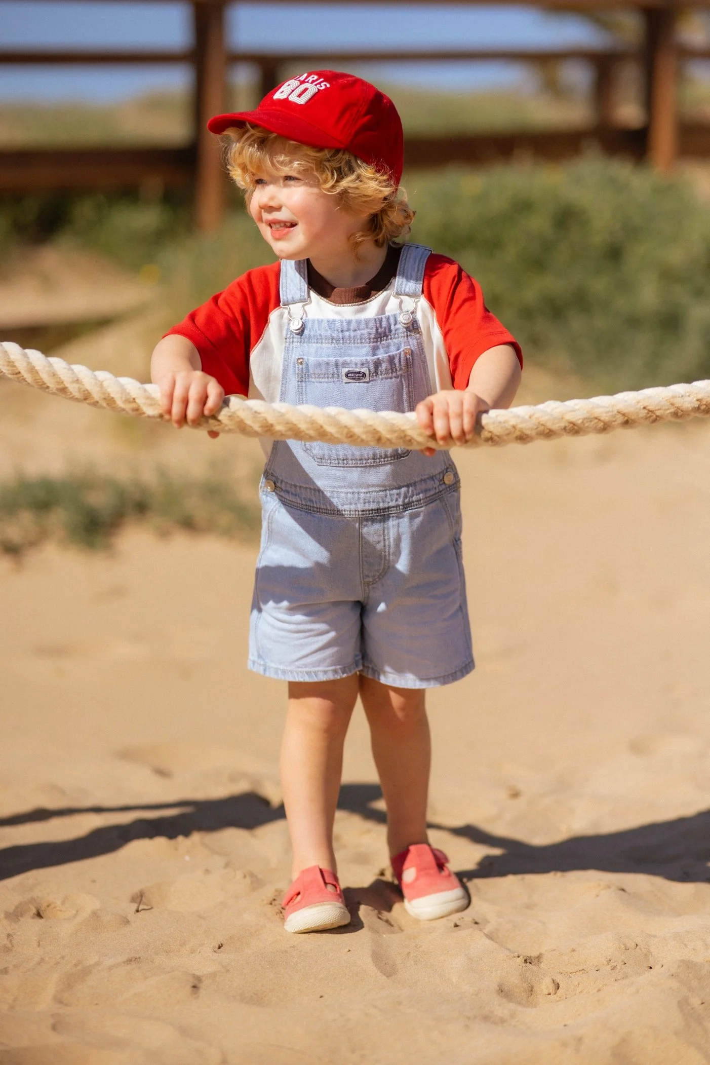 Niño con sombrero rojo, camiseta roja y overoles vaqueros, sosteniendo una cuerda en un parque de juegos al aire libre, sonriente y disfrutando del día.