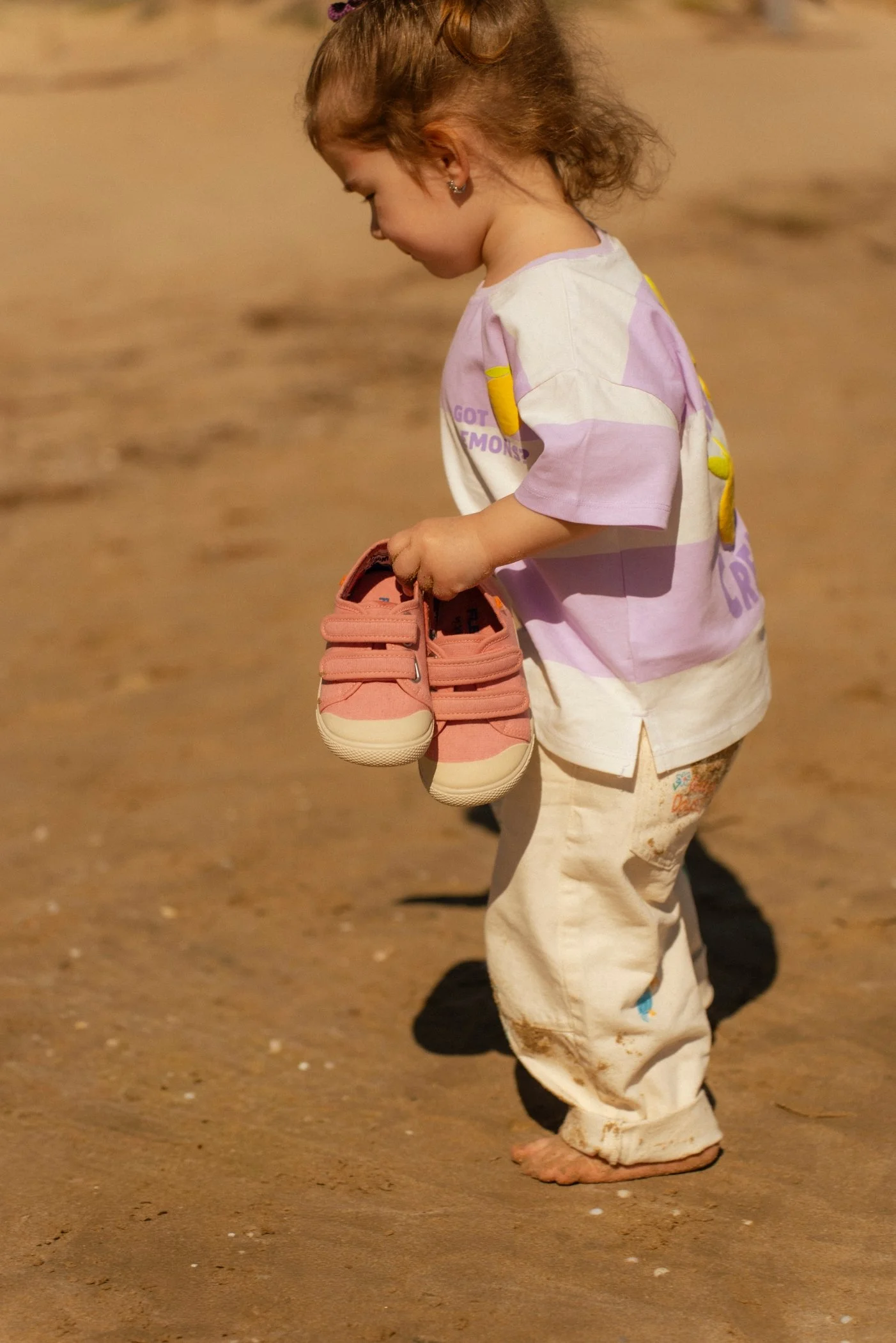 Niña pequeña con ropa clara, de espaldas, sosteniendo unos zapatos rosas en la mano, caminando sobre arena.