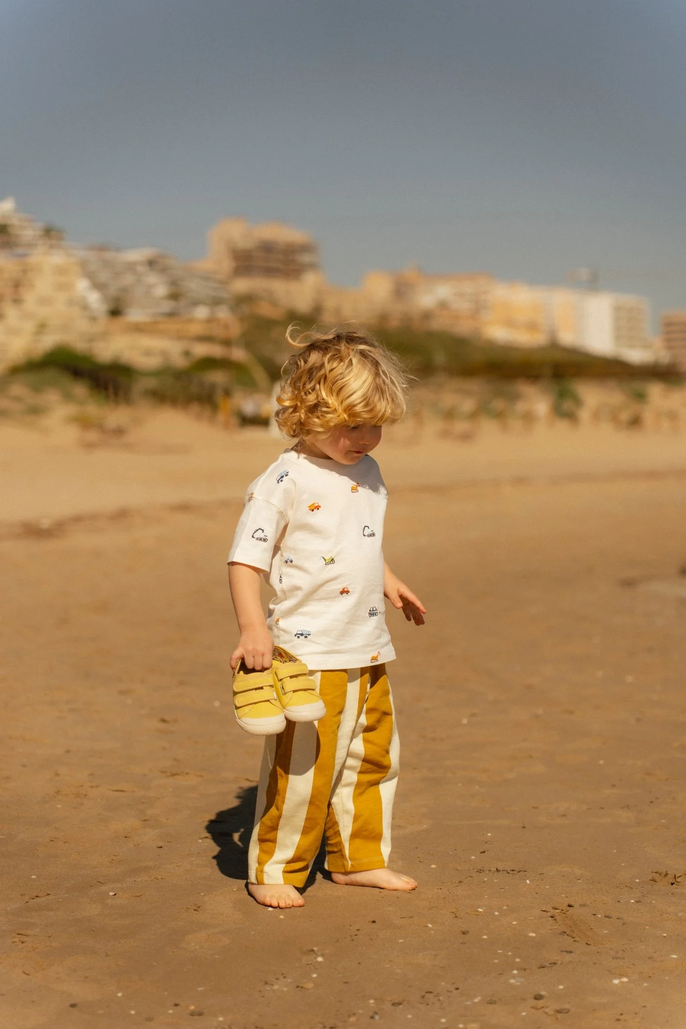 Un niño pequeño con cabello rubio y rizado, de pie en la playa, sosteniendo un par de sandalias amarillas con velcro. Lleva una camiseta blanca con pequeños diseños y pantalones a rayas en tonos amarillos y blancos.