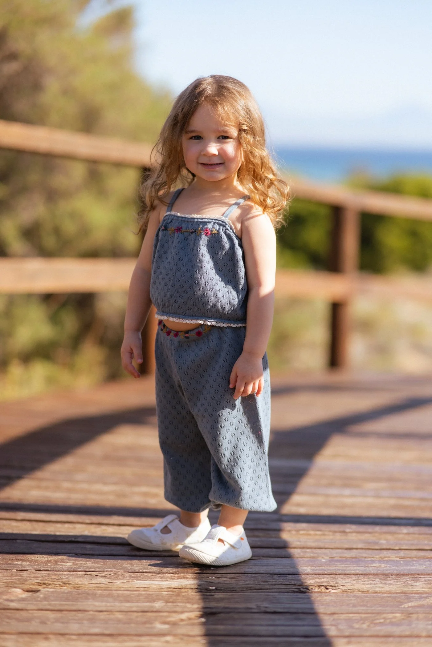 Niña de cabello rizado, con vestido azul y zapatos blancos, en una pasarela de madera con fondo natural y cielo azul.