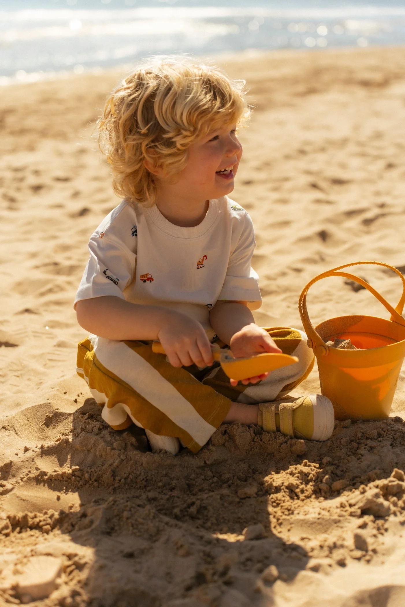 Niño rubio jugando en la arena de la playa, sosteniendo una pala y dientes sonriendo, con un balde naranja junto a él.