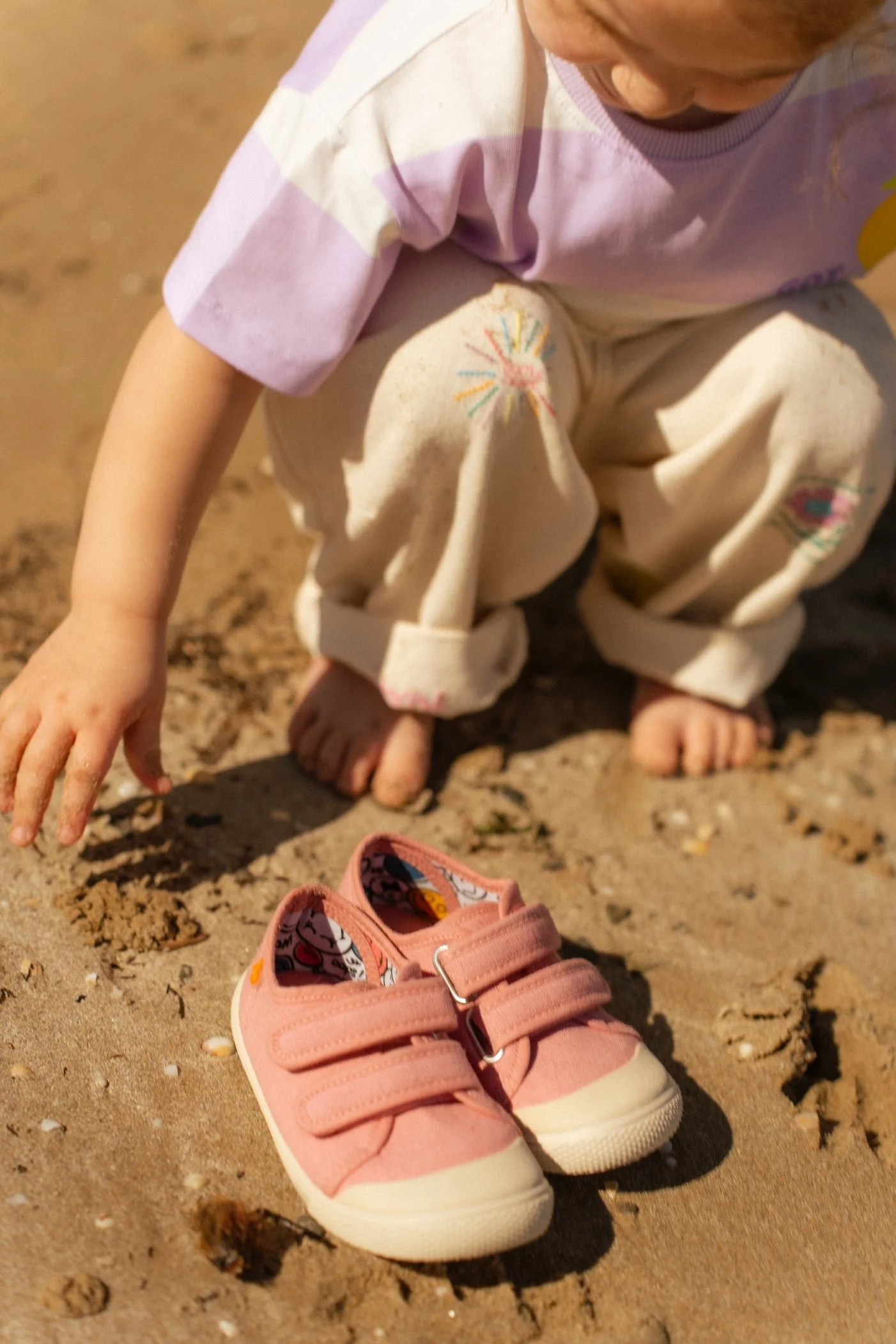 Niña sentada en la arena del mar, con zapatos rosas al frente, y con ropa colorida y pantalones beige con dibujos. Ella está tocando la arena con la mano.