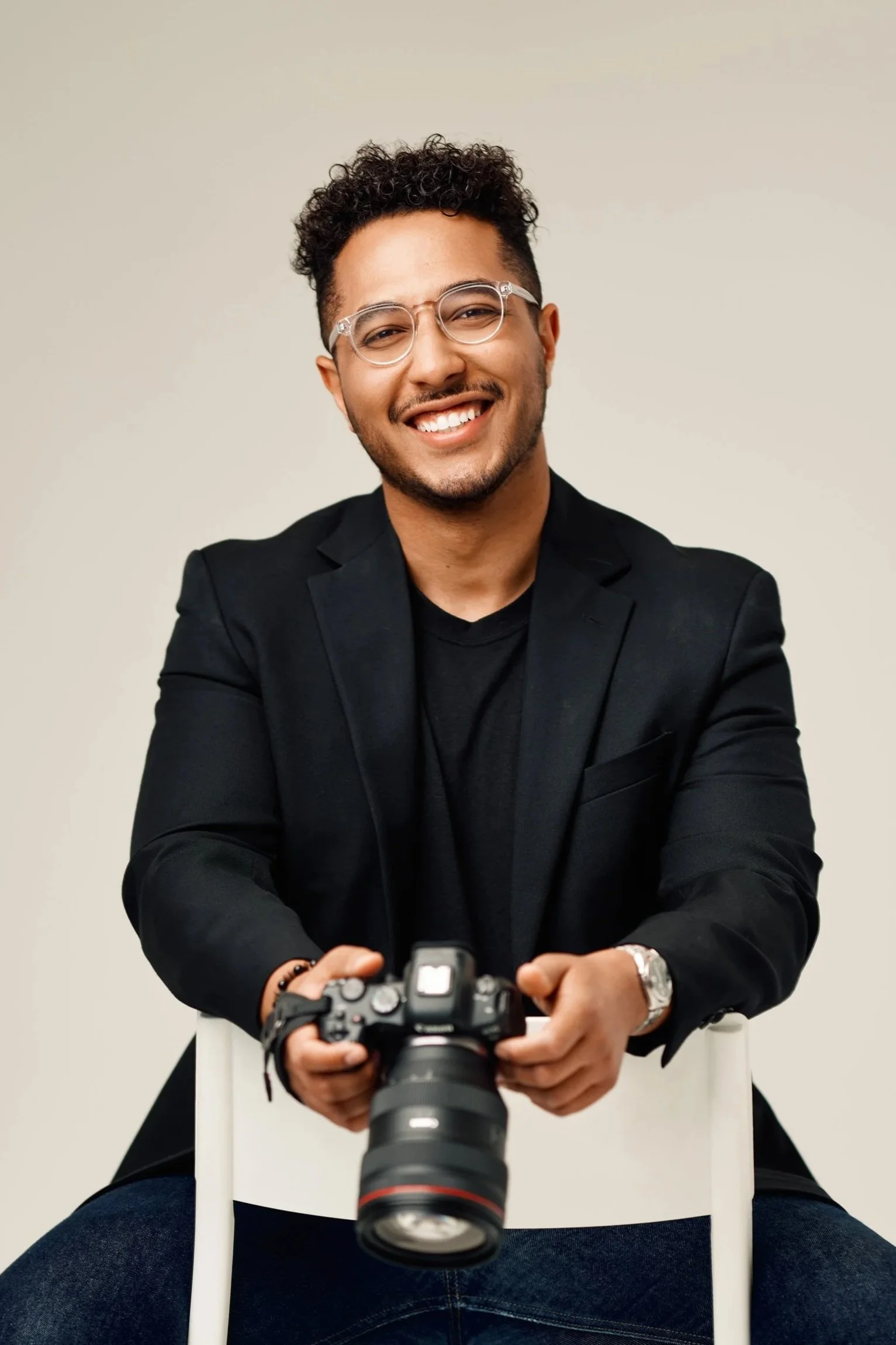 Smiling man with glasses sitting on a chair, holding a camera, wearing a black blazer and dark shirt, against a plain background.