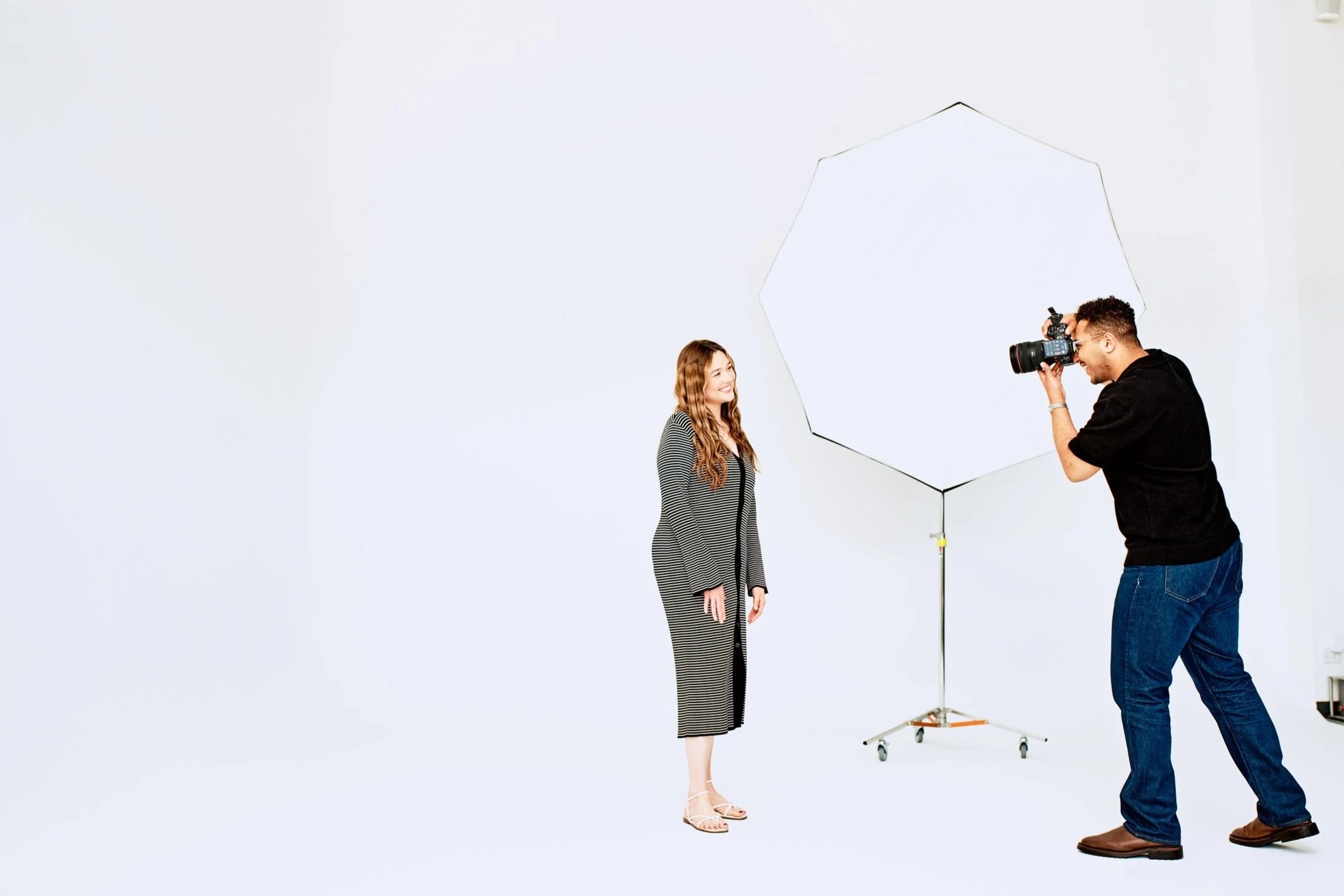 Photographer taking a portrait of a woman in a photography studio with a white backdrop and large octagonal softbox light.