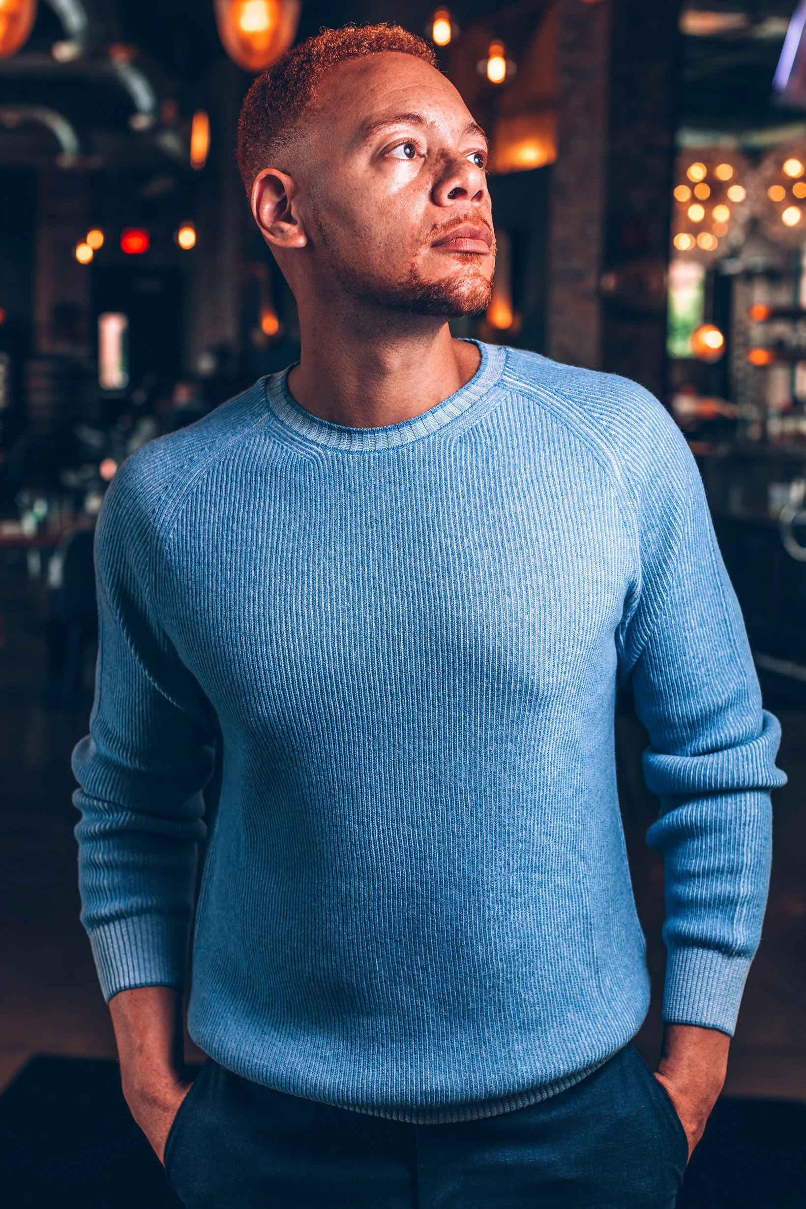 A man wearing a light blue sweater standing in a dimly lit restaurant or bar, looking to the right with hands in his pockets.