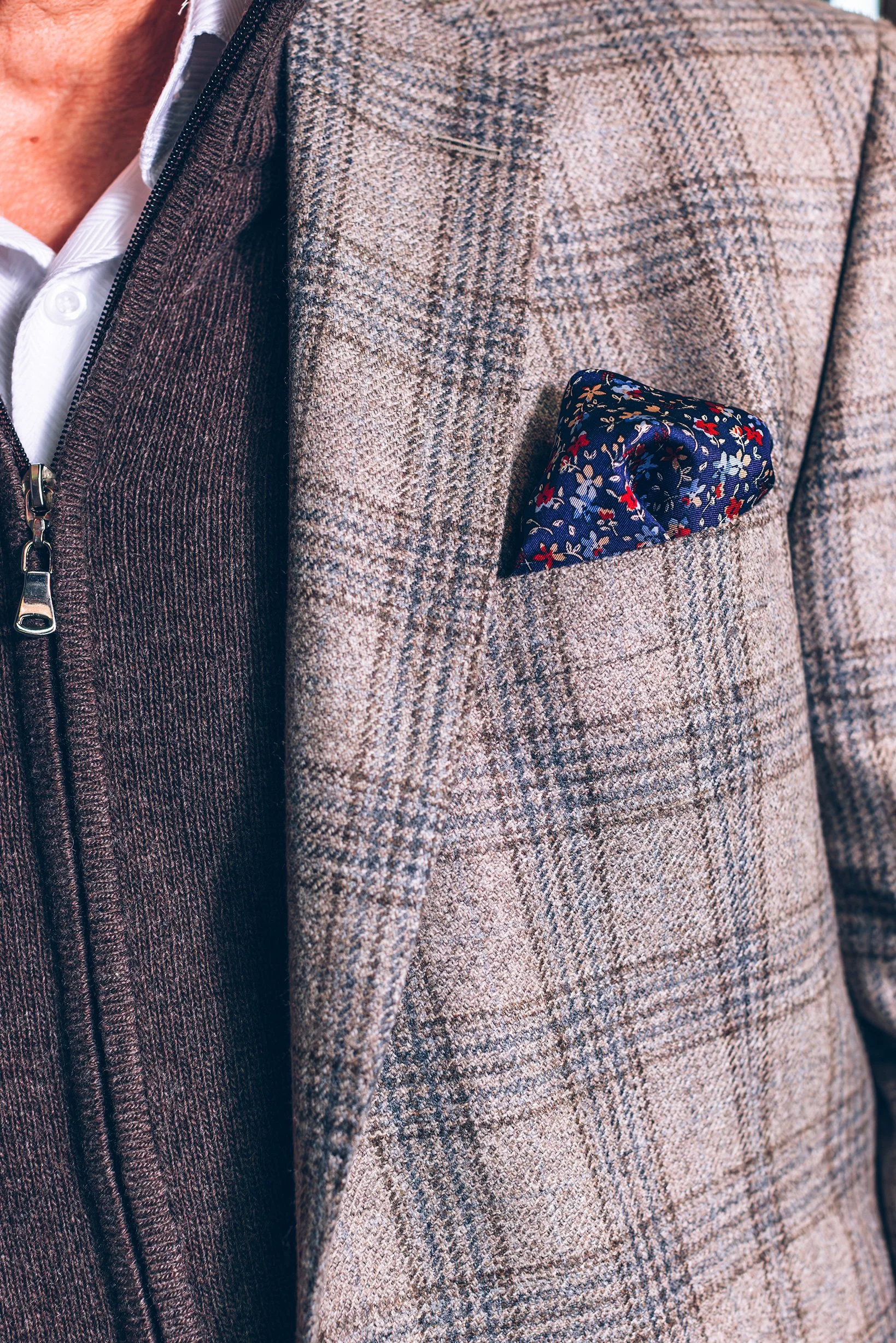 Close-up of a person wearing a checkered beige blazer with a navy floral pocket square and a brown sweater underneath.
